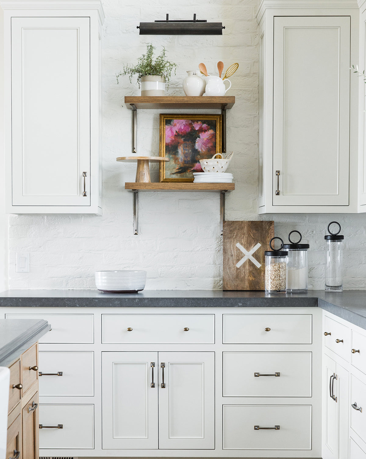 Modern white kitchen with gray countertops, open wooden shelves displaying plants, jars, art, Peonies & Vase for a vintage touch; lower cabinets with silver handles and cereal and utensil containers on the right.