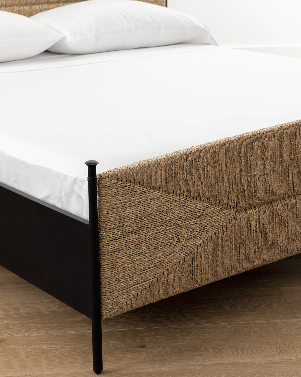 A close-up of the Peregrine Bed featuring a woven seagrass frame with sleek black metal legs, dressed in white sheets and pillows, placed on a wooden floor by McGee & Co.