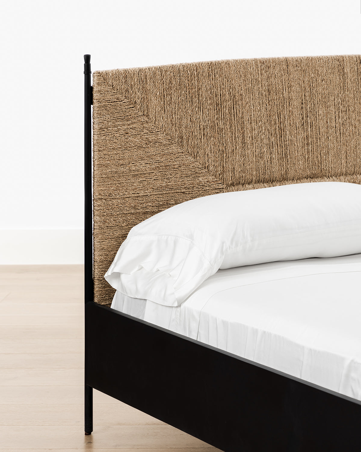 Close-up of the Peregrine Bed with a woven seagrass headboard in a diagonal pattern, sleek black metal frame, crisp white sheets, and light wooden flooring, by McGee & Co.