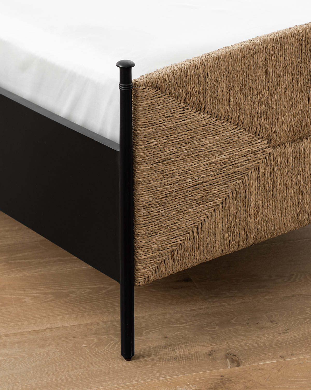 Close-up of the Peregrine Bed’s black metal frame and woven seagrass headboard at the corner, shown on a light wood floor with a white mattress on top, by McGee & Co.
