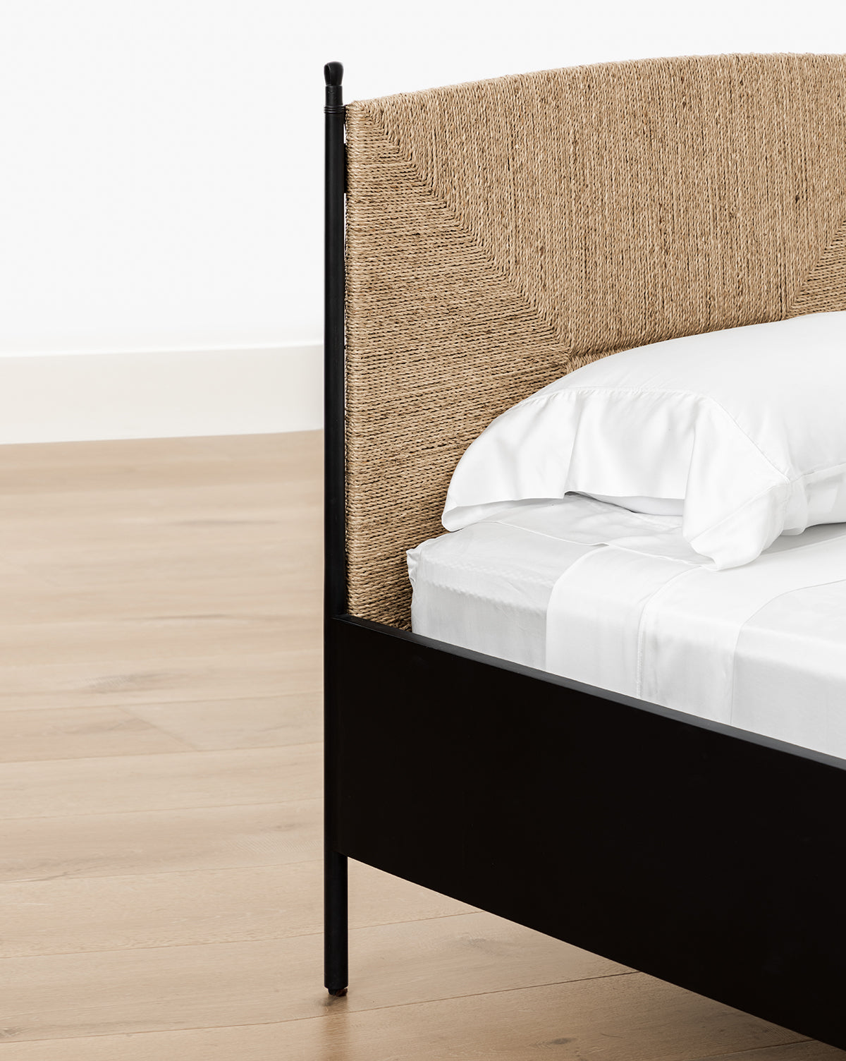 Close-up of the Peregrine Bed featuring a black metal frame and woven seagrass headboard. It is dressed in white sheets and pillows, placed on a light wooden floor against a white wall, by McGee & Co.