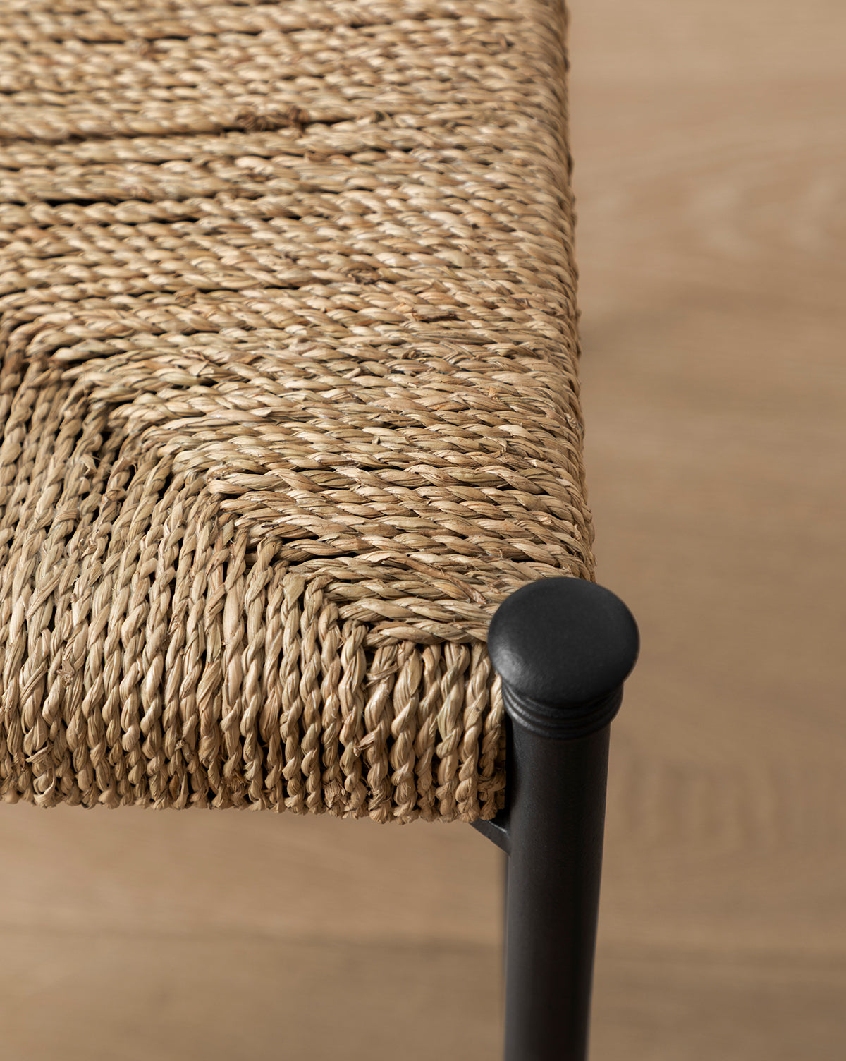Close-up of the Peregrine Chair’s woven seagrass seat on a black metal frame, highlighting its detailed texture and rounded corner joint. Set against a light wood floor, it’s ideal for enhancing your dining chair collection with style, by McGee & Co.