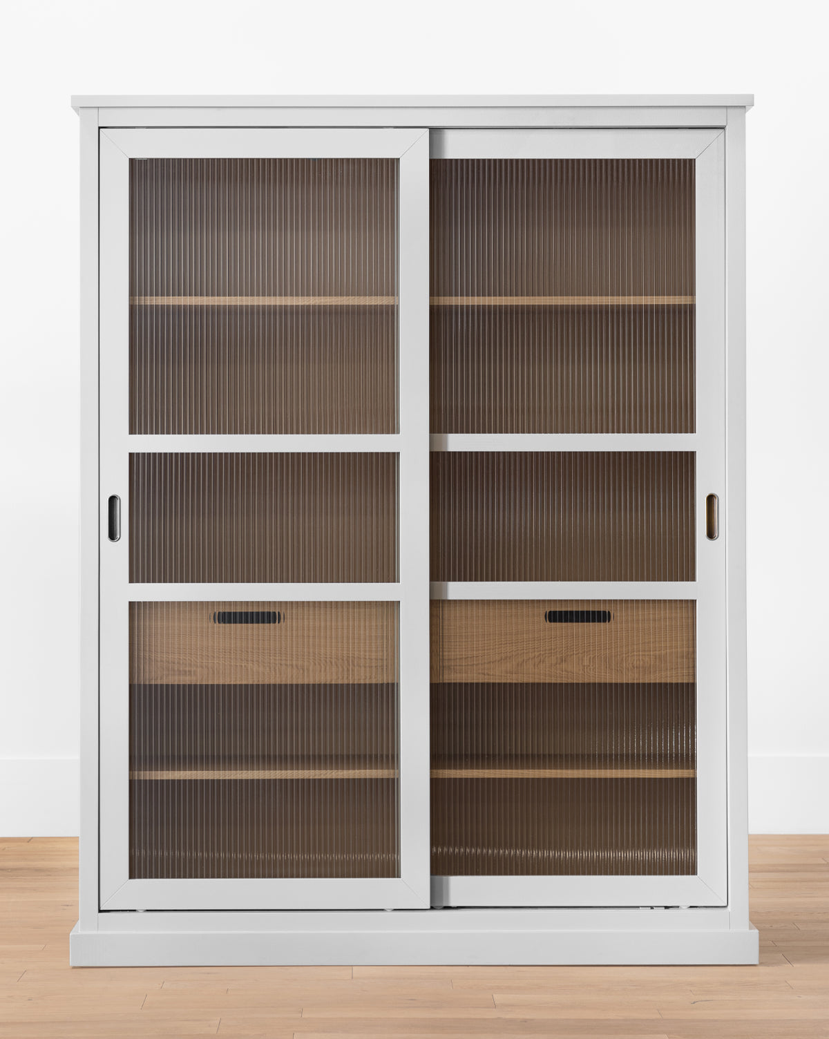 The Phillips Cabinet features painted oak with reeded glass doors, wooden shelves, and two drawers inside. Antique brass handles add charm as it stands on a light wood floor against a white wall, by McGee & Co.