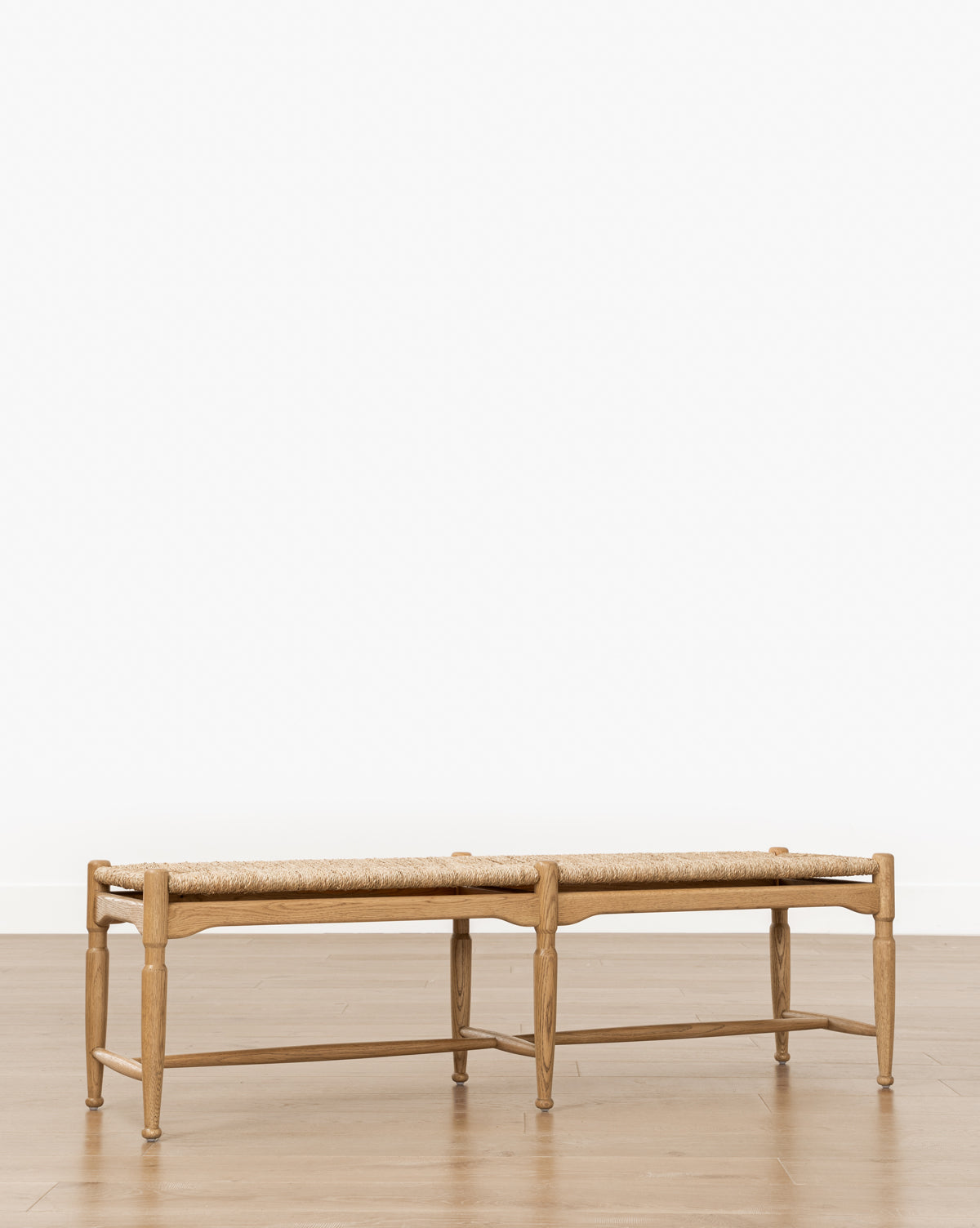 The Phipps Woven Bench, featuring a seagrass seat and natural wood frame, sits on a wooden floor against a plain white wall, by McGee & Co.