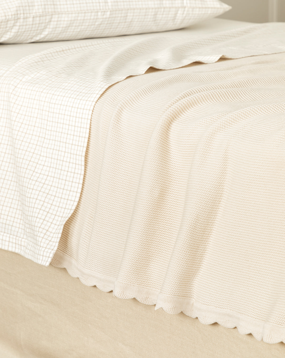 A neatly made bed featuring the Pinnacle Knit Kids Blanket in cream over a checked white sheet and pillowcase, creating a cozy, minimalist look with soft, neutral tones.