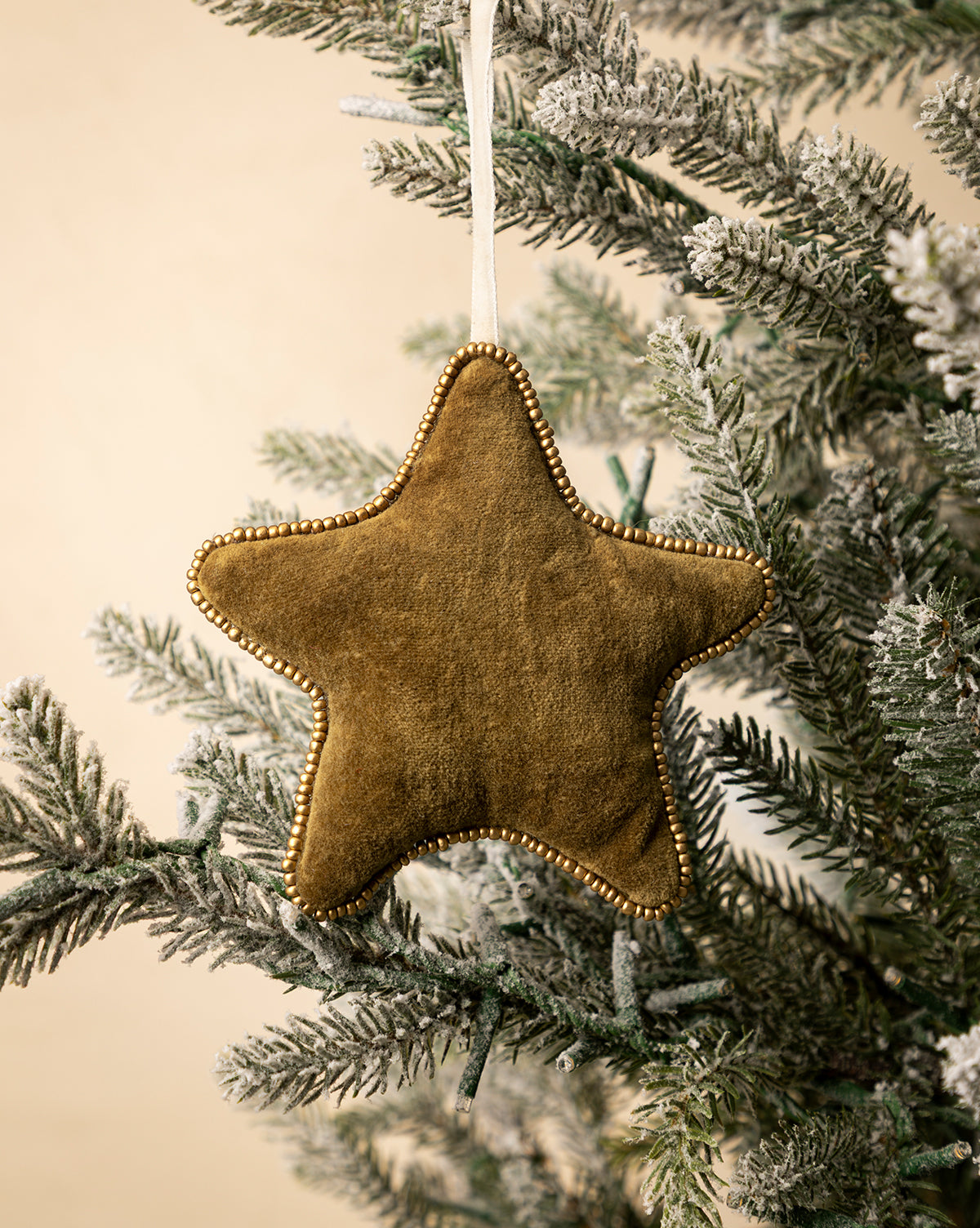 The Plush Beaded Star Ornament, adorned with gold beads and festive design, hangs from a snowy holiday tree branch against a light beige background, by McGee & Co.