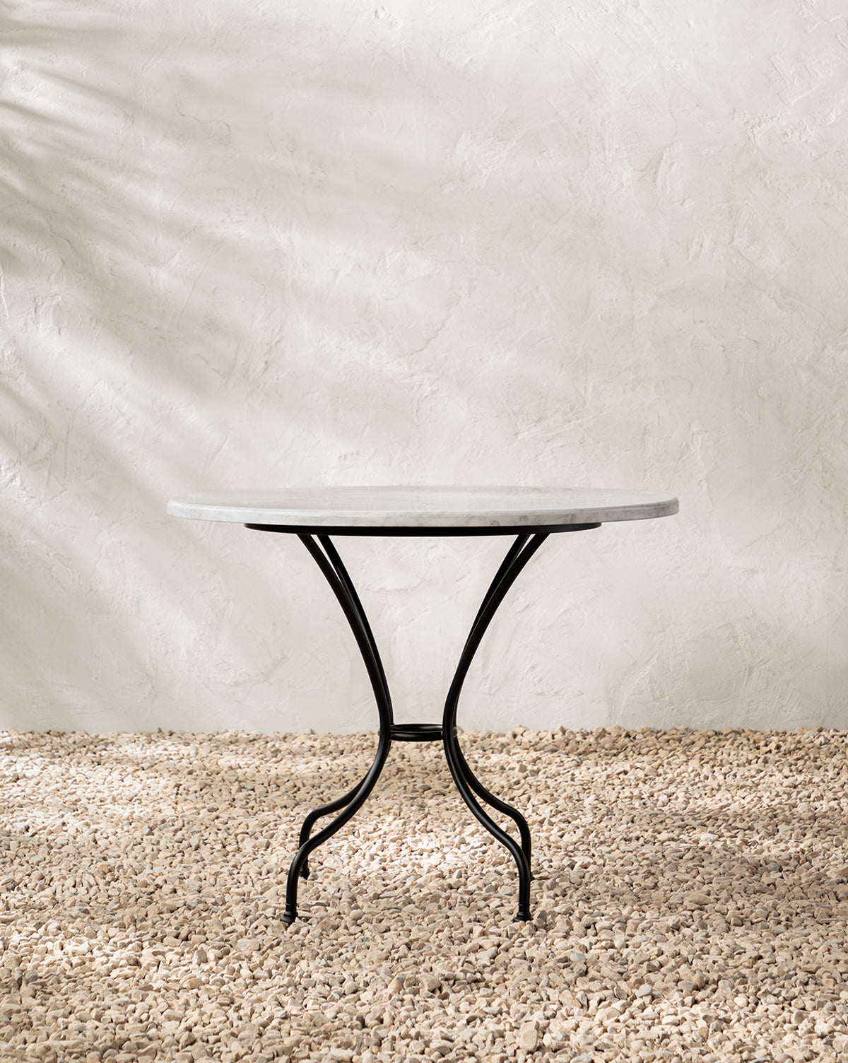 The Pomeroy Outdoor Bistro Table features a sleek white marble round tabletop and black metal legs, standing on a textured pebble floor against a light, textured wall with soft shadows by McGee & Co.