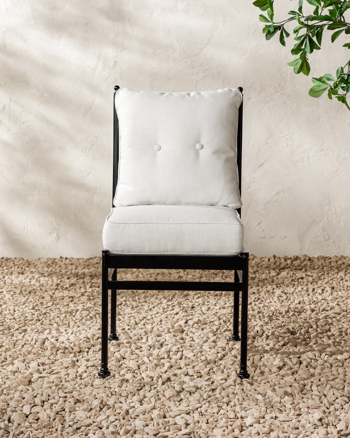 The Pomeroy Outdoor Dining Chair, featuring a black powder-coated steel frame and white performance fabric cushion, sits on a textured light stone floor with a leafy green plant partially visible against a neutral wall, by McGee & Co.