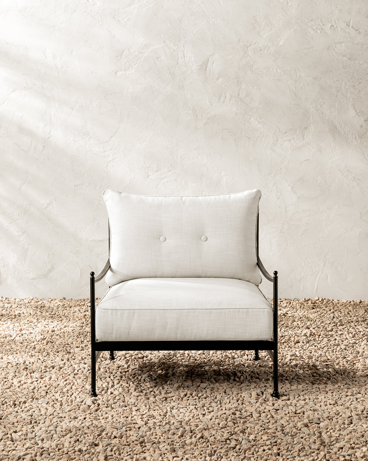 The Pomeroy Outdoor Lounge Chair has a powder-coated steel frame and off-white performance fabric cushions, shown on a textured light brown pebble floor with a white plaster wall backdrop, by McGee & Co.
