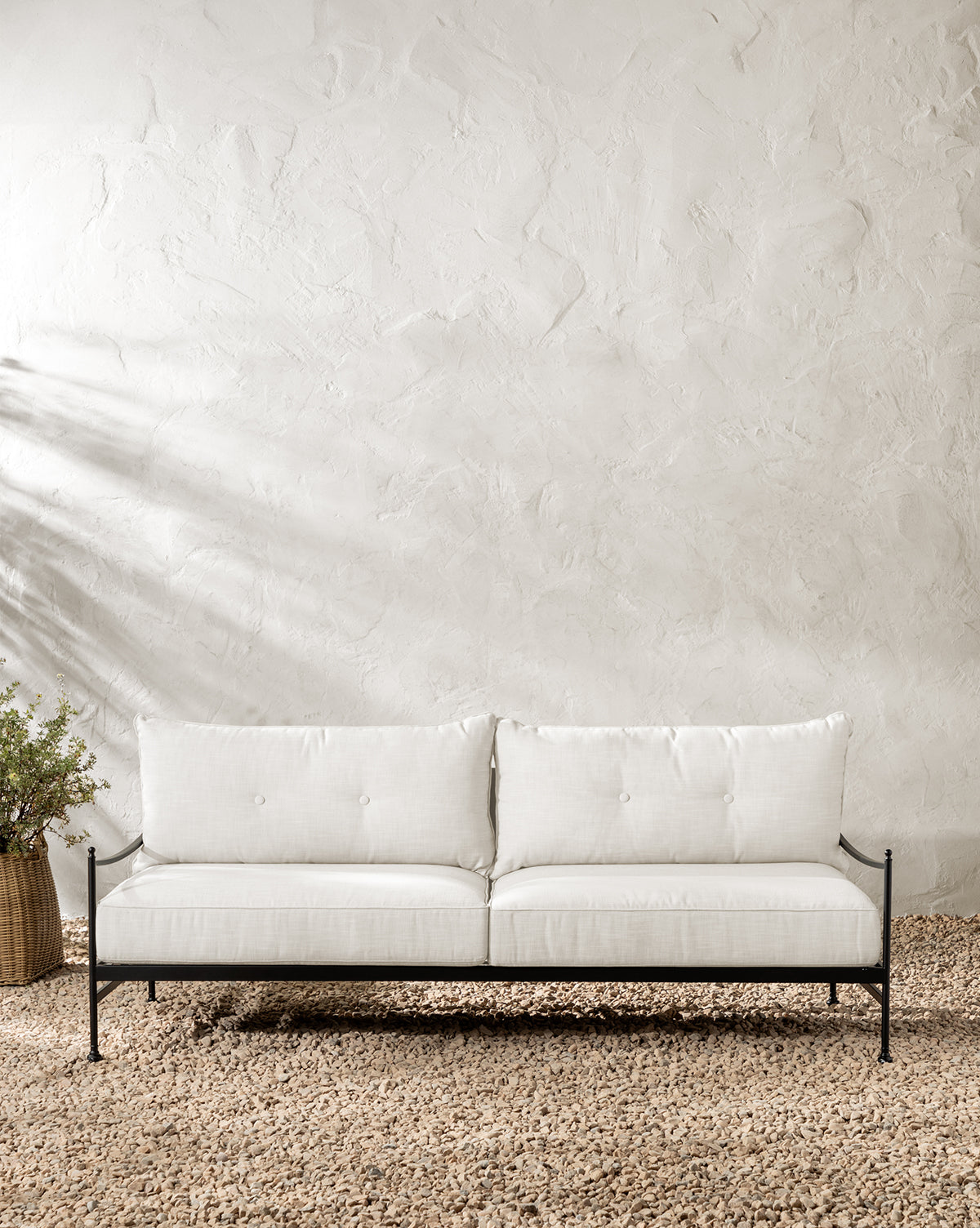 A modern white Pomeroy Outdoor Sofa 83 with slim black powder-coated steel legs sits on a pebble floor by a textured beige wall, accompanied on the left by a woven basket containing a leafy plant, by McGee & Co.