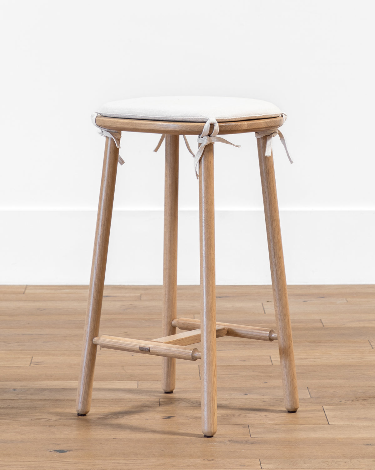 The Ralph Oak Counter Stool features four legs and a circular white linen cushion with ties, shown on a light wood floor against a plain white wall—perfect for your kitchen counter, by McGee & Co.