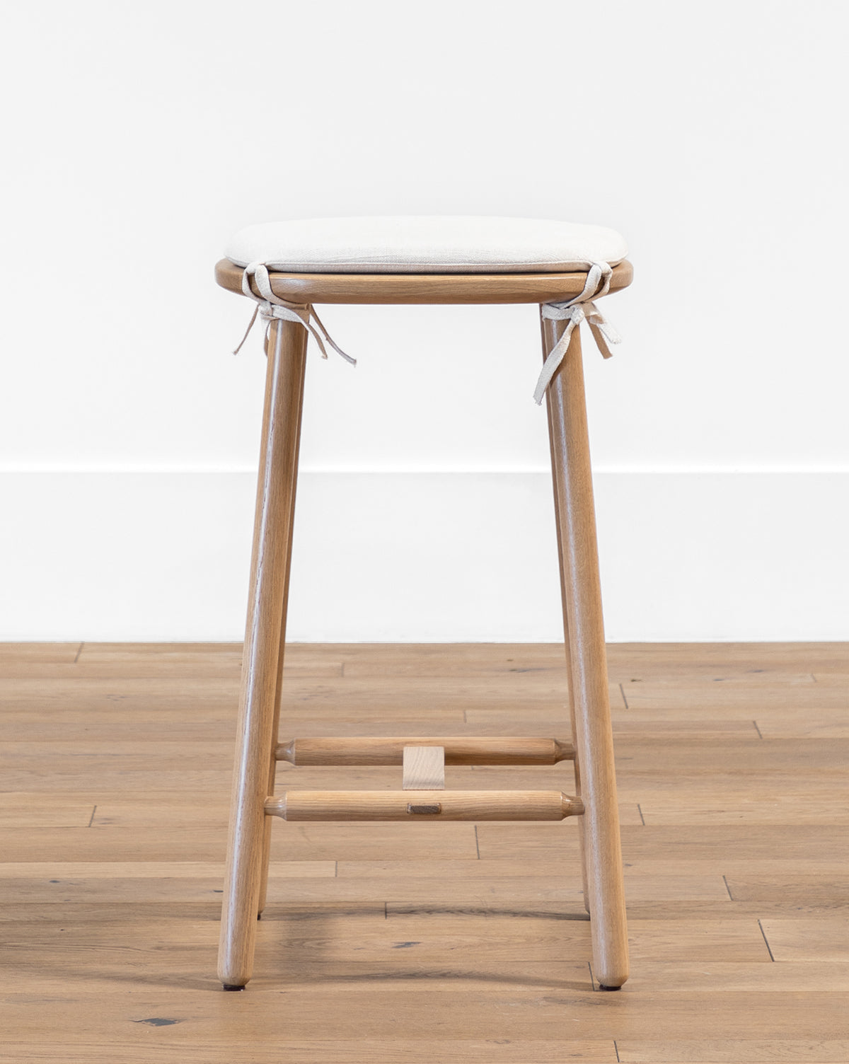 The Ralph Oak Counter Stool features a wooden oak frame and a light linen cushion tied at the corners, placed on a wooden floor against a white wall by McGee & Co.