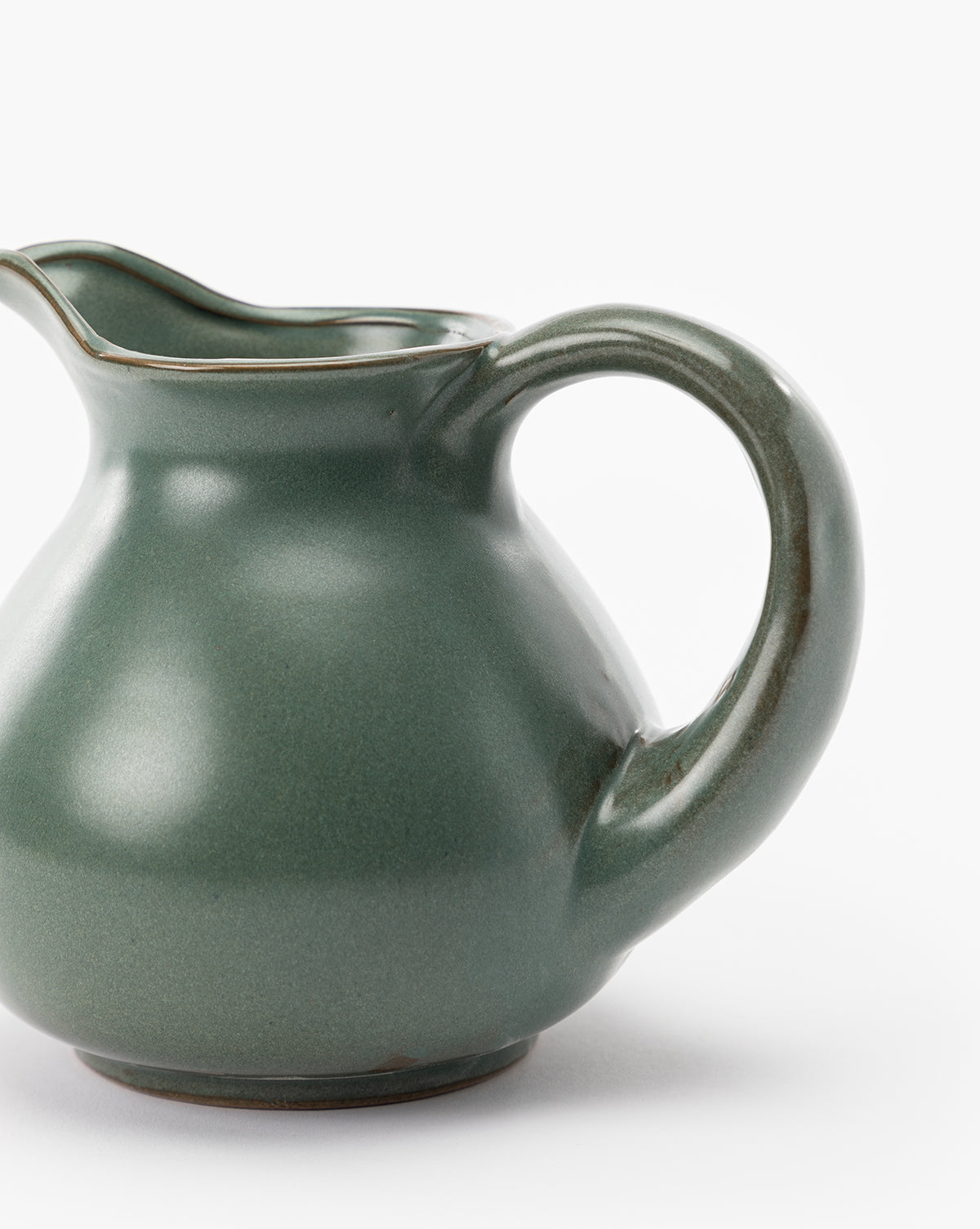 A close-up of the Rayen Stoneware Pitcher with a matte green glaze, rounded body, curved handle, and small spout, set against a plain white background, by McGee & Co.