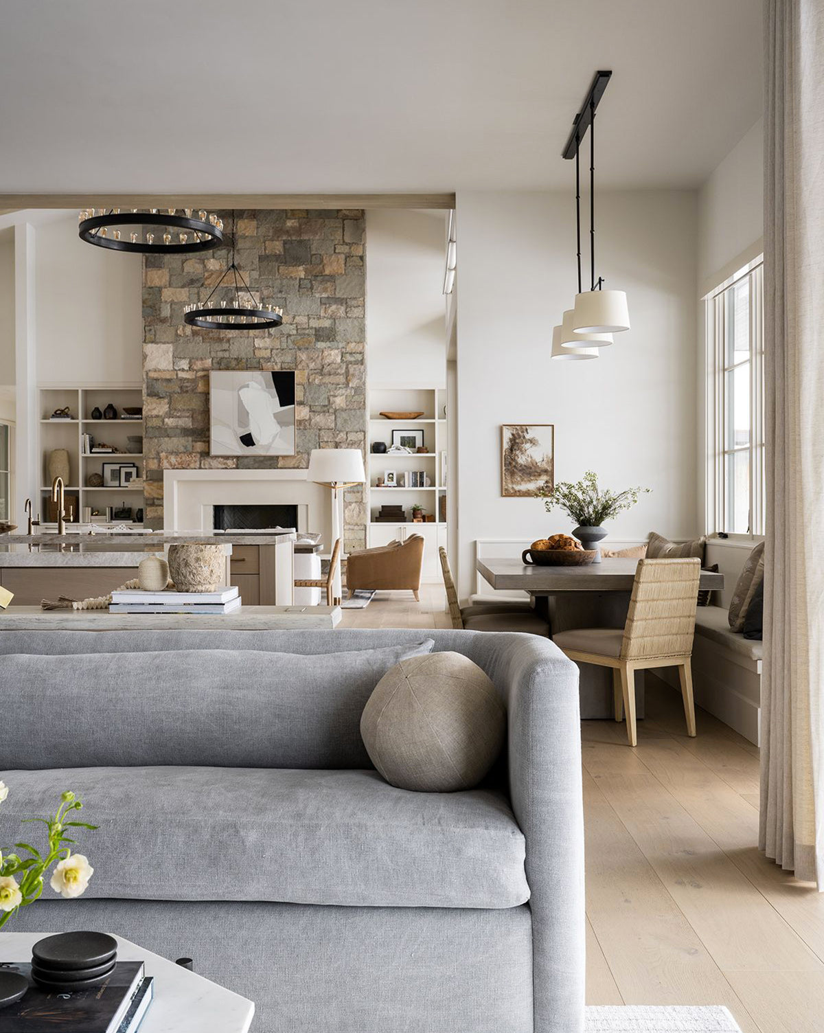 A modern, bright living area showcases a Reese Sofa (84-96) with down-wrapped cushions, a stone fireplace, handcrafted alder wood shelves in the kitchen, a round dining table with chairs, and large windows bringing in natural light by McGee & Co.