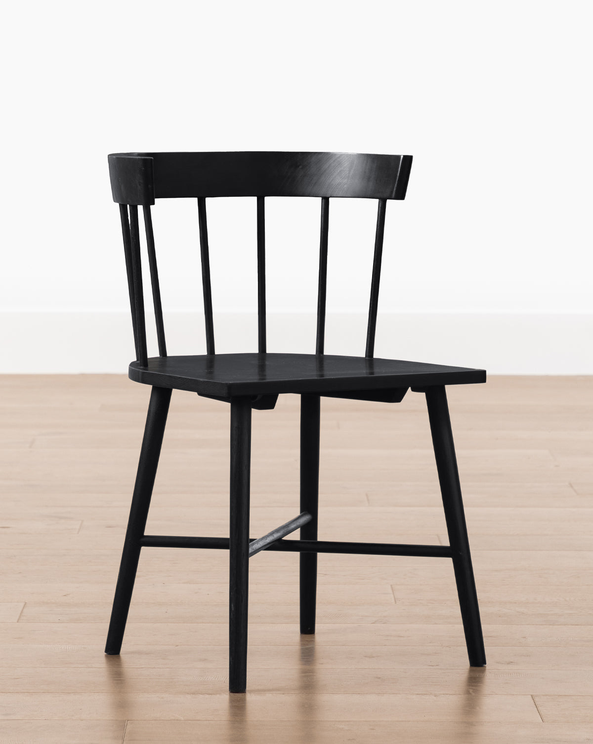 A Reeves Dining Chair, with a curved backrest and vertical slats, angled legs, and a flat seat, sits on a light wood floor beside a plain white wall by McGee & Co.