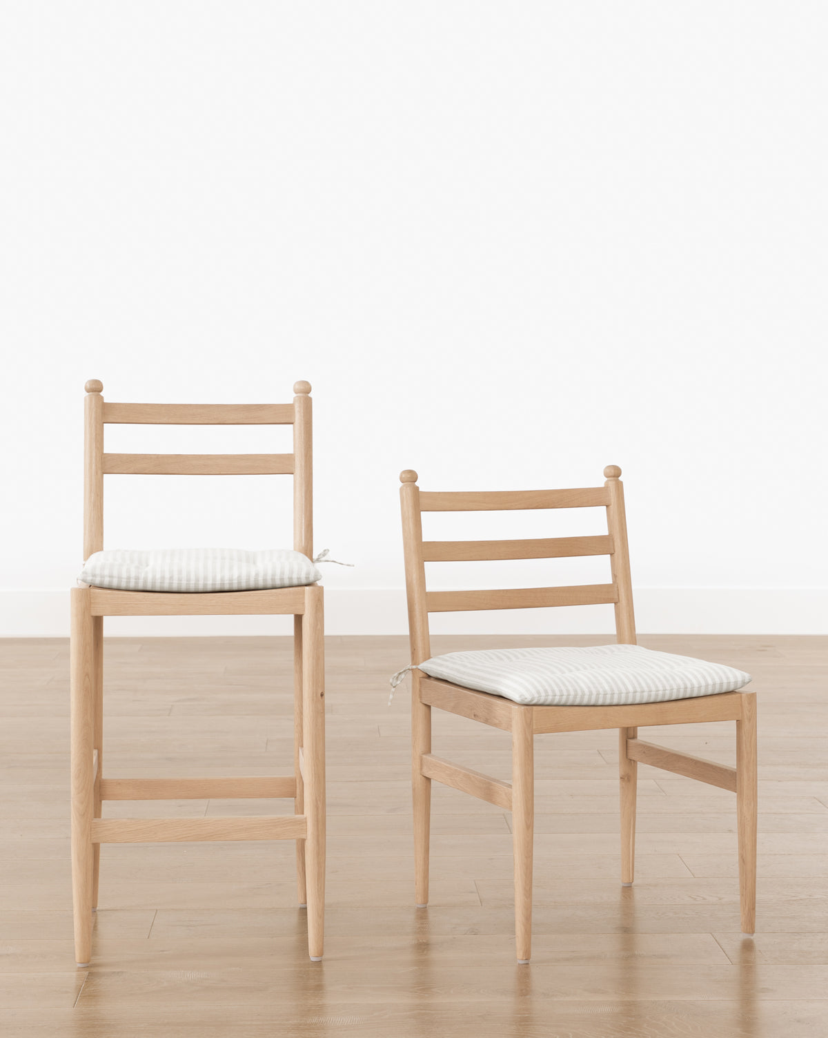 A Romany Counter Stool and a matching white oak dining chair, both with striped linen cushions and ladder backs, sit side by side on a wood floor. Their simple, modern design adds style to any space by McGee & Co.