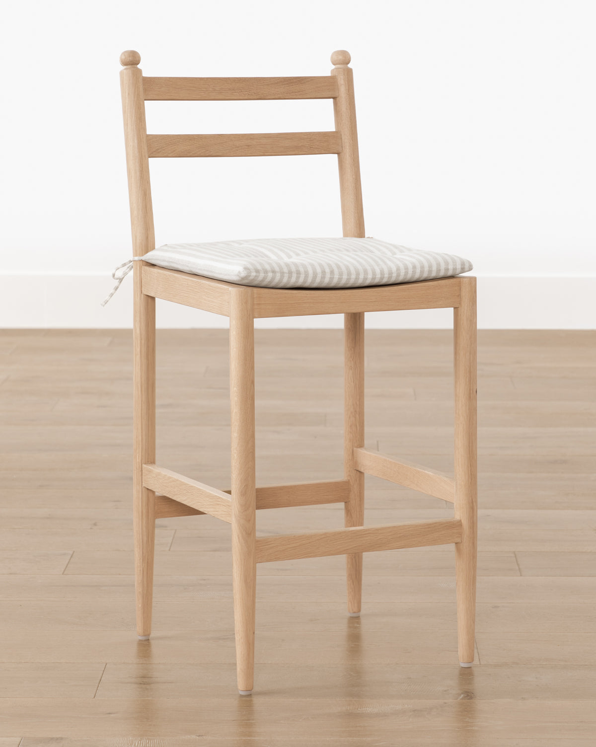 The Romany Counter Stool features a white oak frame and a striped linen cushion tied to the backrest, set on a wood floor against a plain white background, by McGee & Co.