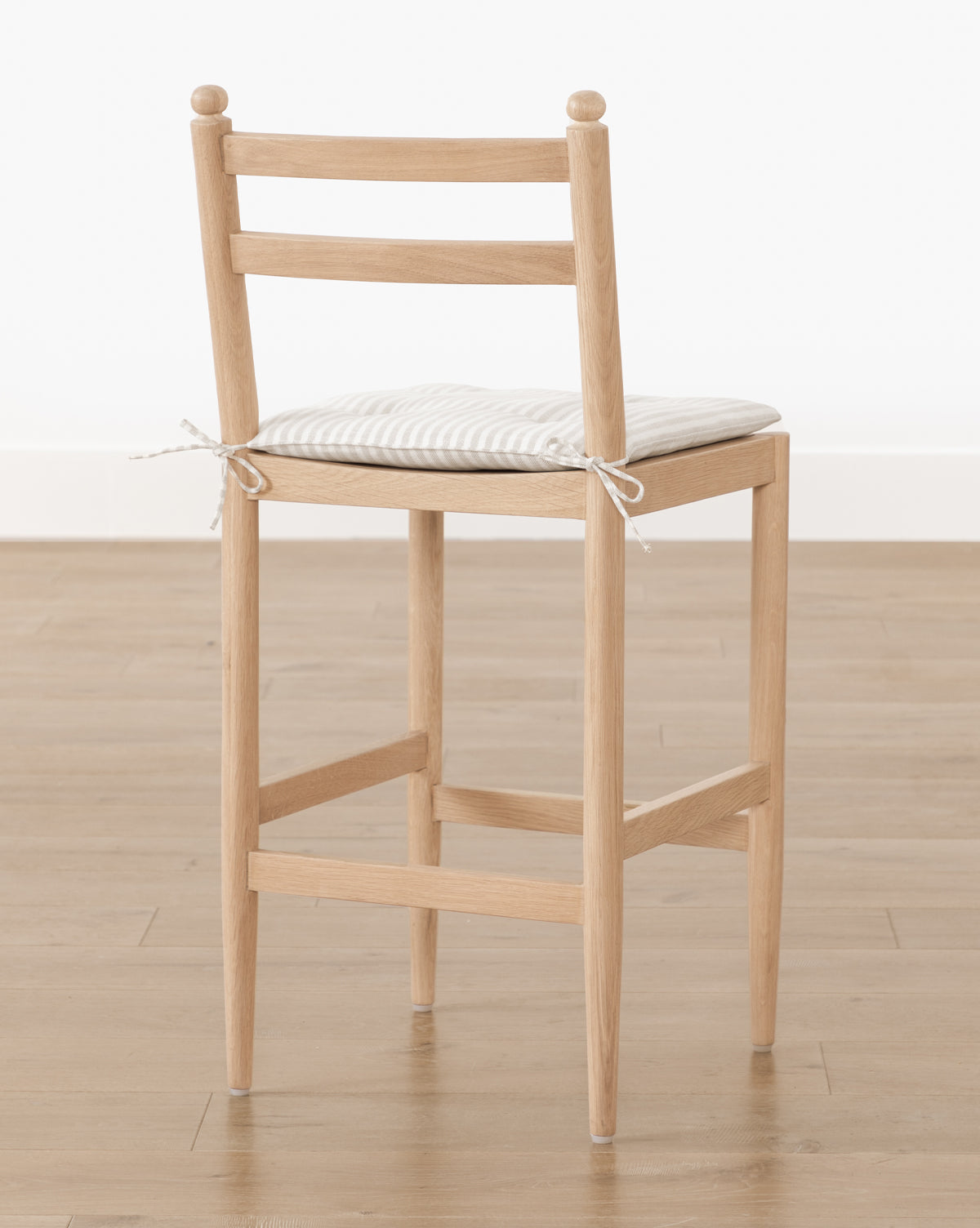 The Romany Counter Stool, featuring a light white oak frame and striped linen tie-on cushion, sits on a wooden floor against a plain white wall, by McGee & Co.