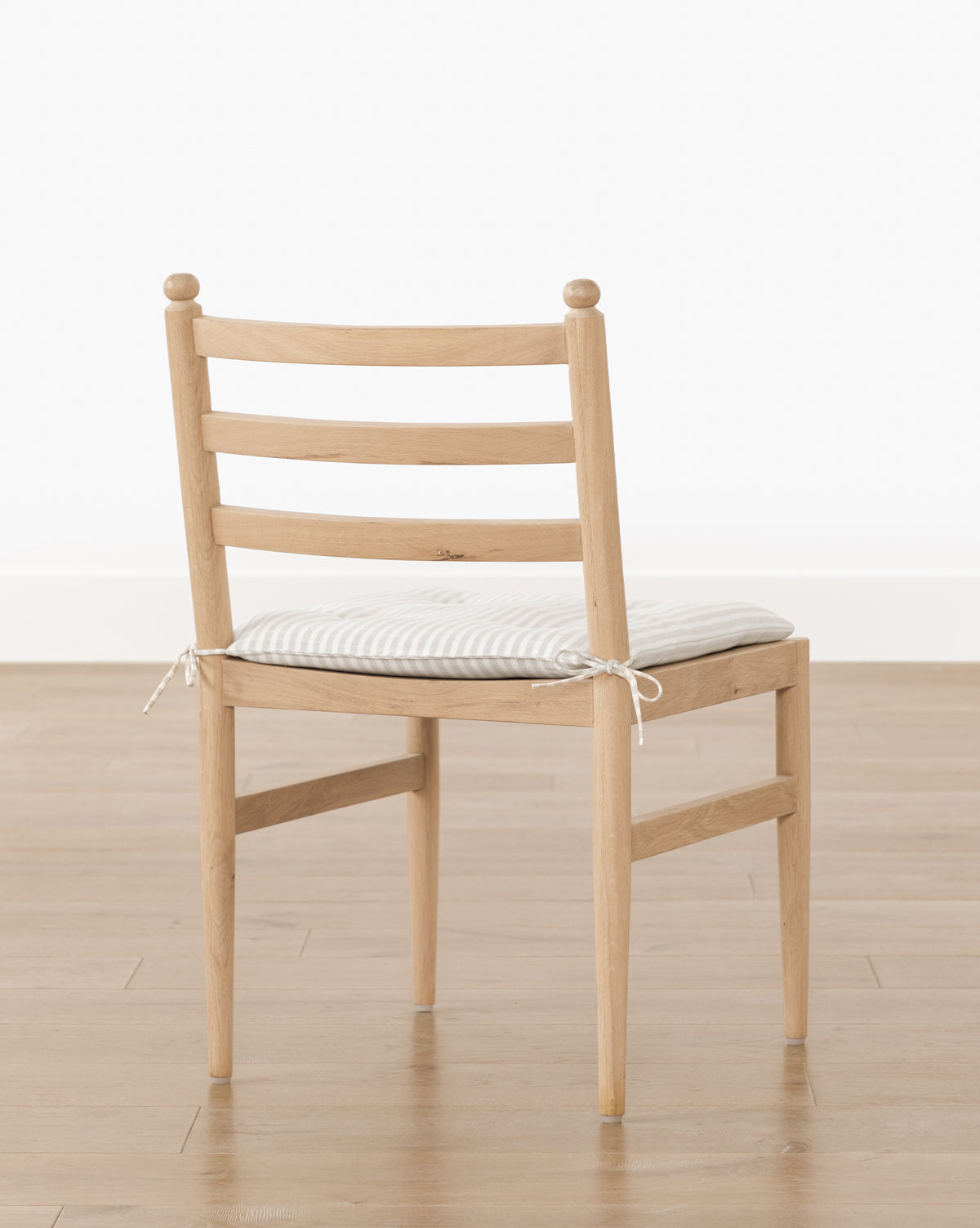 The Romany Dining Chair, crafted from white oak, has a ladder-style back and tapered legs. It includes a light-striped linen cushion secured to the seat. Shown on a wooden floor with a plain white wall backdrop by McGee & Co.