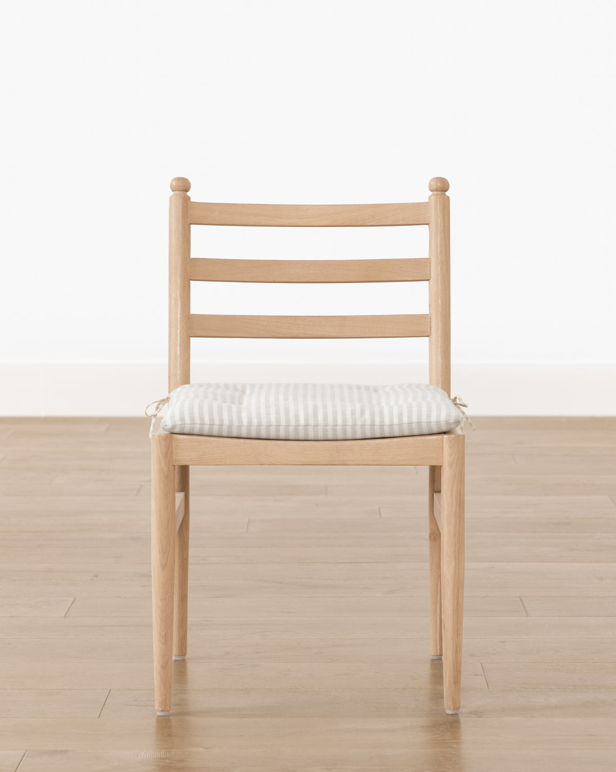 The Romany Dining Chair features a white oak frame, ladder-back design, and a light checkered linen cushion, set on a wooden floor against a plain white wall by McGee & Co.
