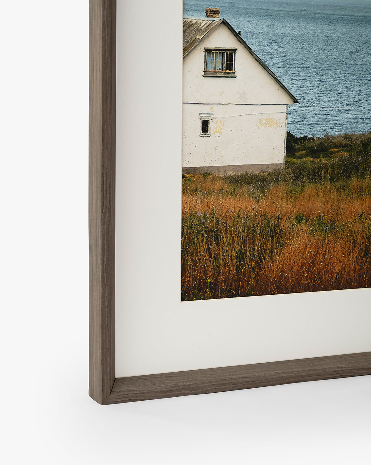 Room with a View: A gallery-quality, framed photograph capturing the weathered corner of a white house, grassy field, and blue water beyond. Only part of the house is shown, enhanced by a dark walnut frame, by McGee & Co.