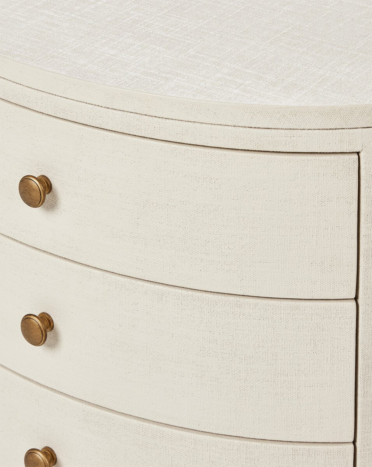 Close-up of the Rosalie Nightstand, an ivory-painted piece with a linen texture, three curved drawers with round brass knobs, and a smooth rounded top edge. This elegant nightstand provides stylish and practical bedside storage by McGee & Co.