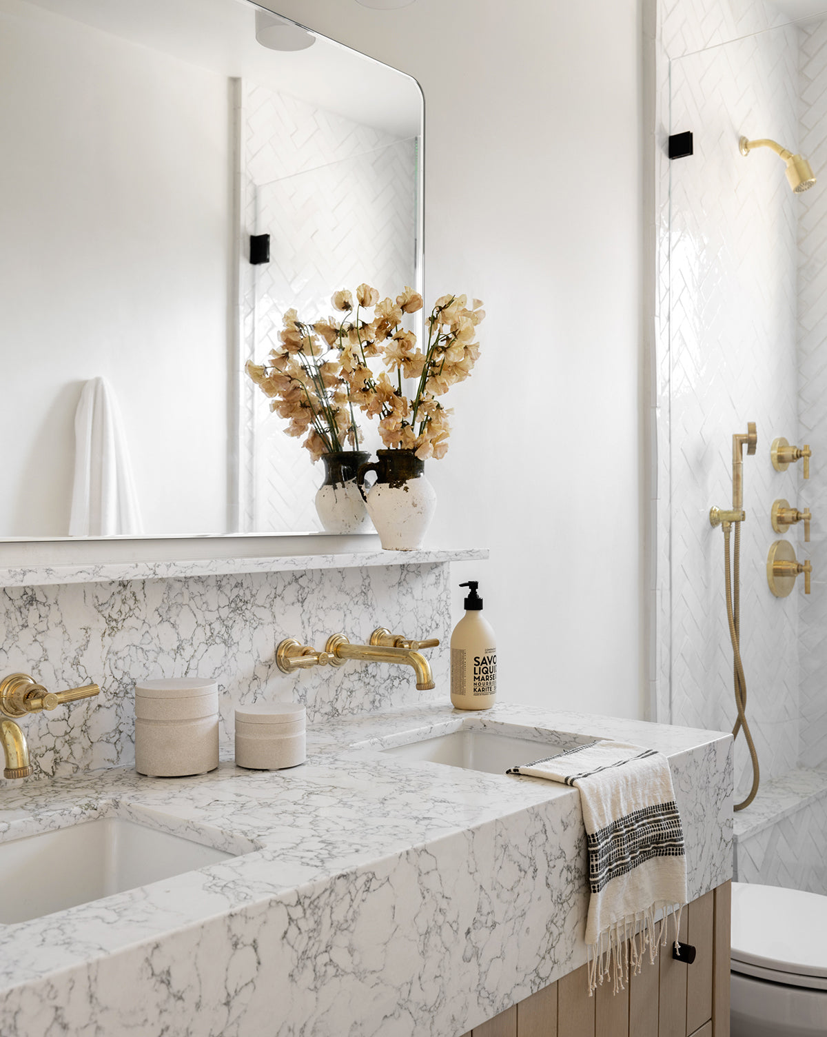 A modern bathroom with a marble countertop, gold fixtures, dried flowers, and a large mirror. The shower has white tile and gold hardware. A towel, soap bottle, and the Barton Sandstone Canister decorate the counter.