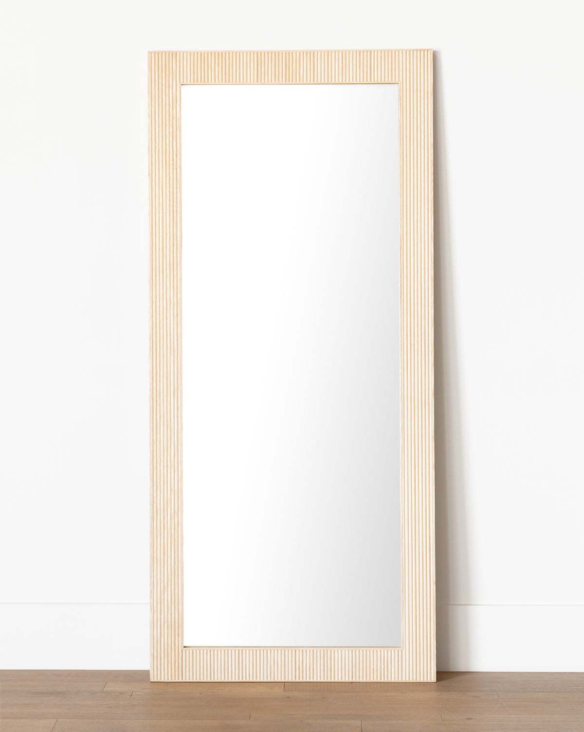 The Rowan Floor Mirror is a tall, rectangular piece with a ribbed oak frame featuring vertical grooves, leaning against a white wall on a wooden floor, by McGee & Co.