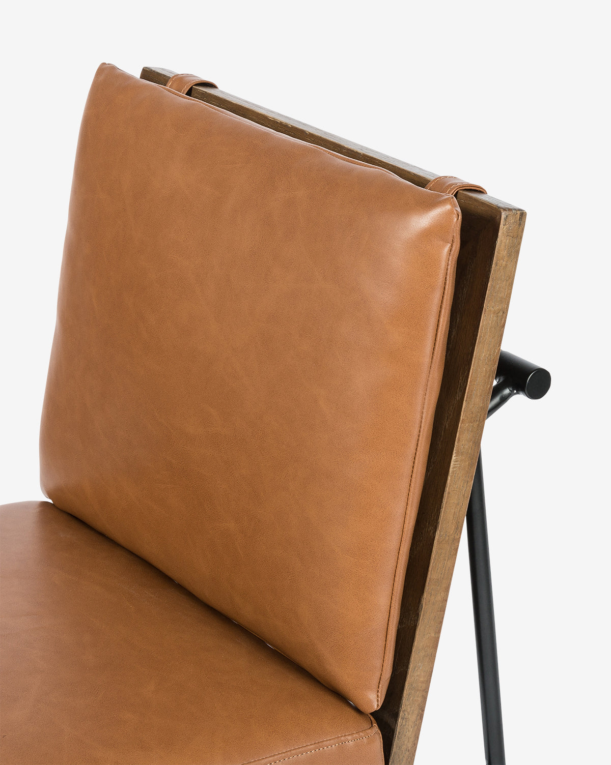 A close-up of the Selas Dining Chair shows its wooden frame, tan leather backrest and seat cushions, and black iron legs. The chair features a modern, minimalist design, by McGee & Co.