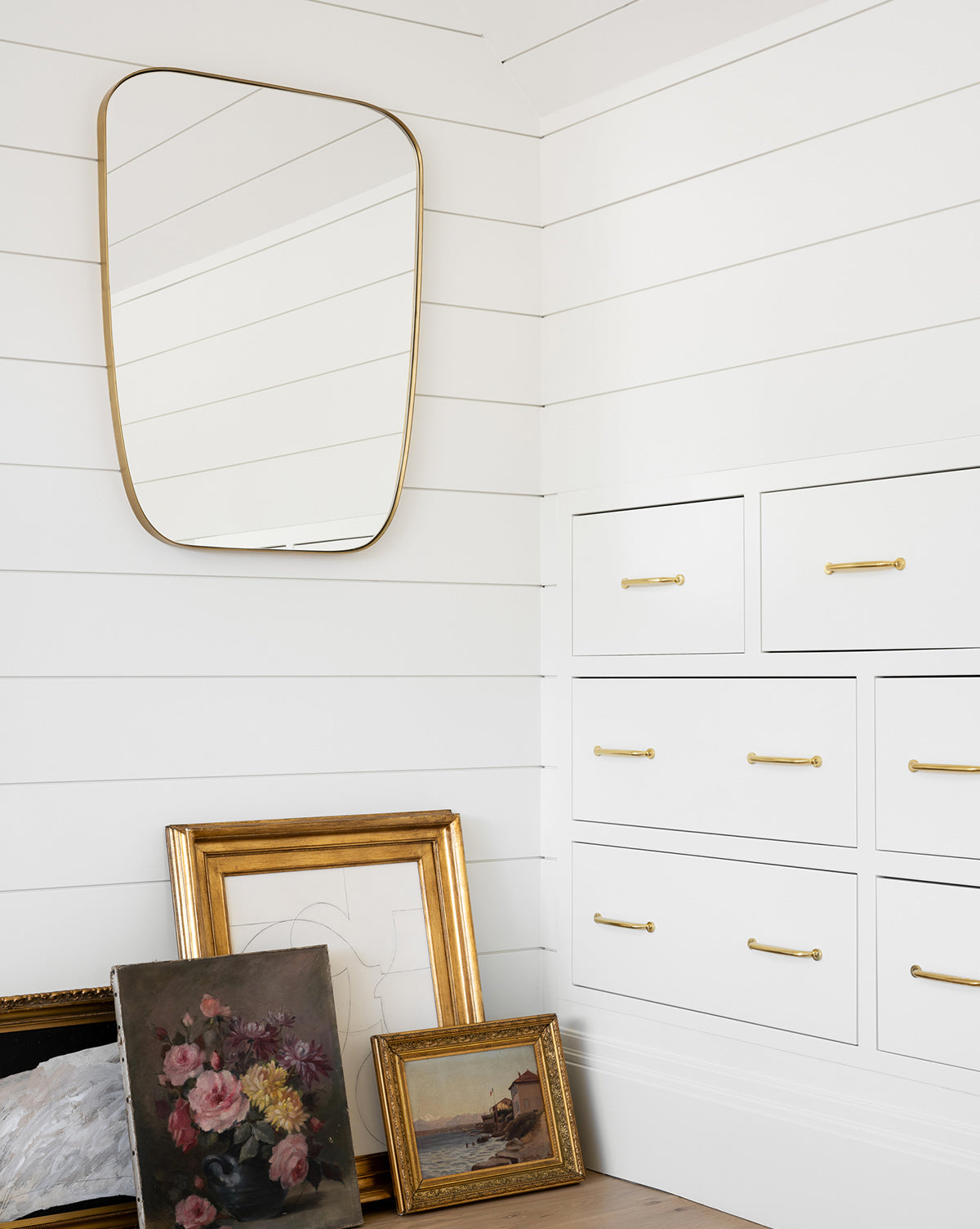 A white shiplap room features a Jace Inset Mirror, white drawers with gold handles, and framed wall art, including a floral painting, leaning against the wall by McGee & Co.