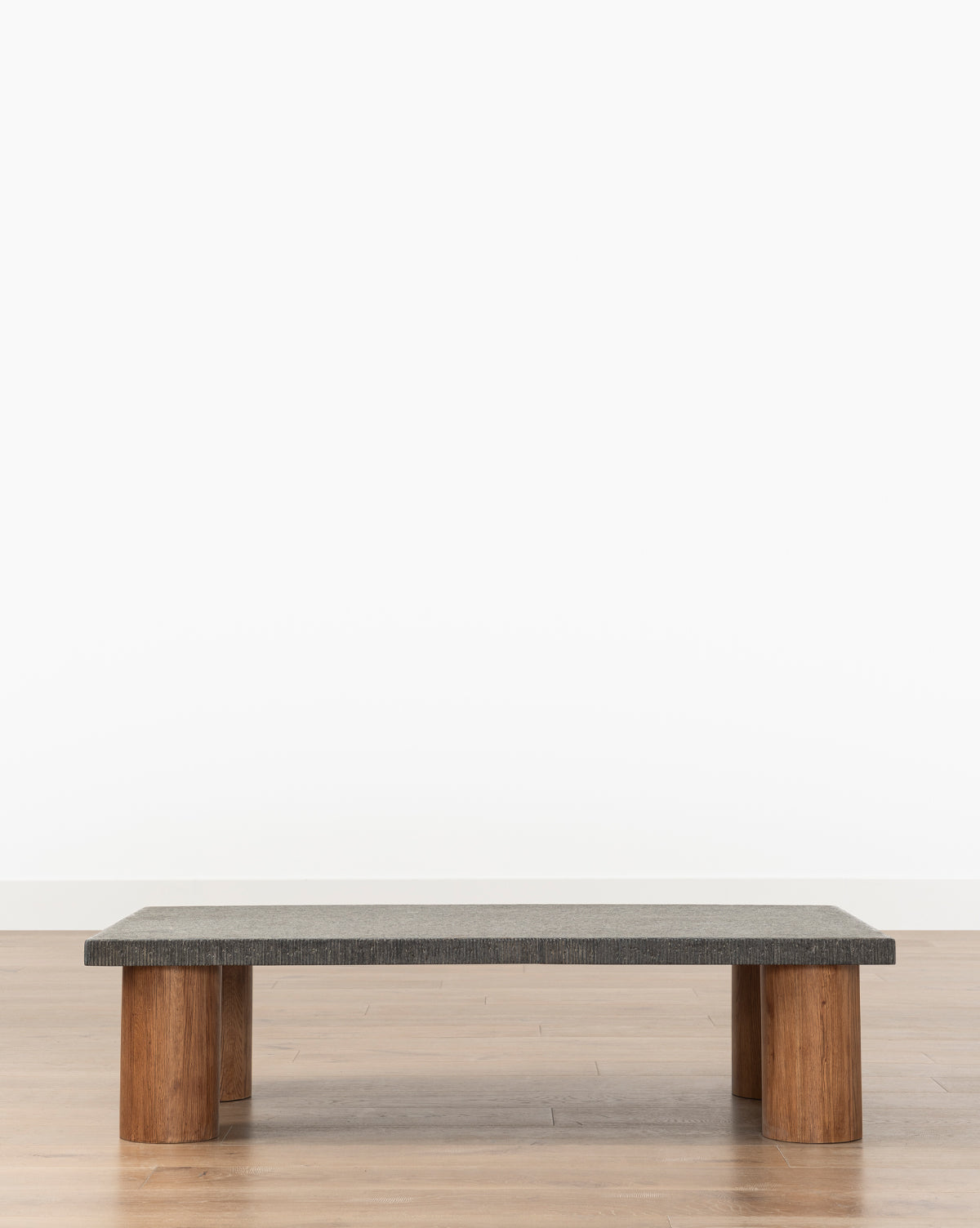 The Sinclair Coffee Table boasts a vintage-inspired design with a rectangular dark granite top and four thick cylindrical wooden legs, displayed on a light wooden floor against a plain white background, by McGee & Co.