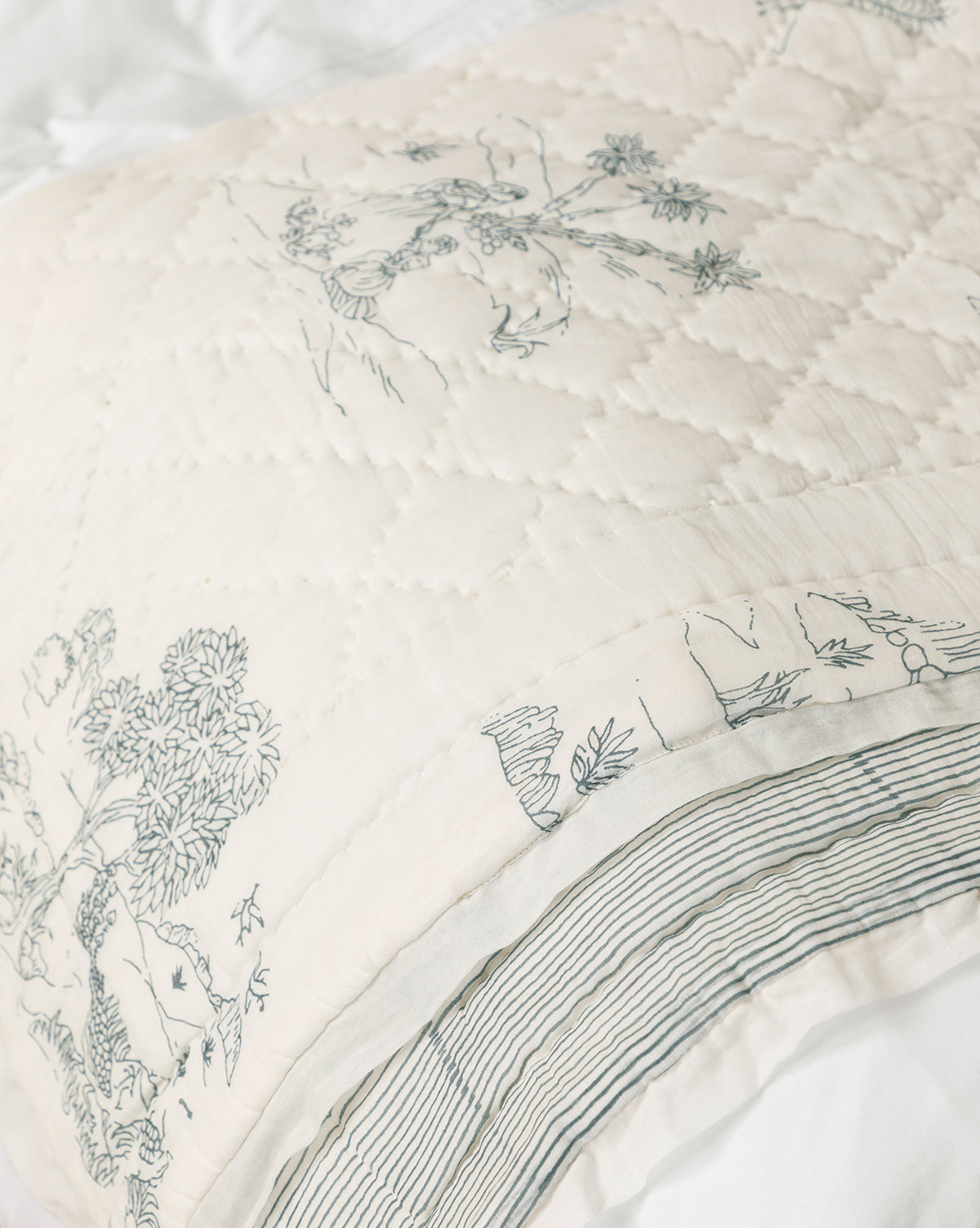A close-up of the Solano Block Print Kids Quilt shows cream cotton bedding with quilted stitching and blue line art of trees and landscapes, finished with subtle blue stripes and a printed border, by McGee & Co.