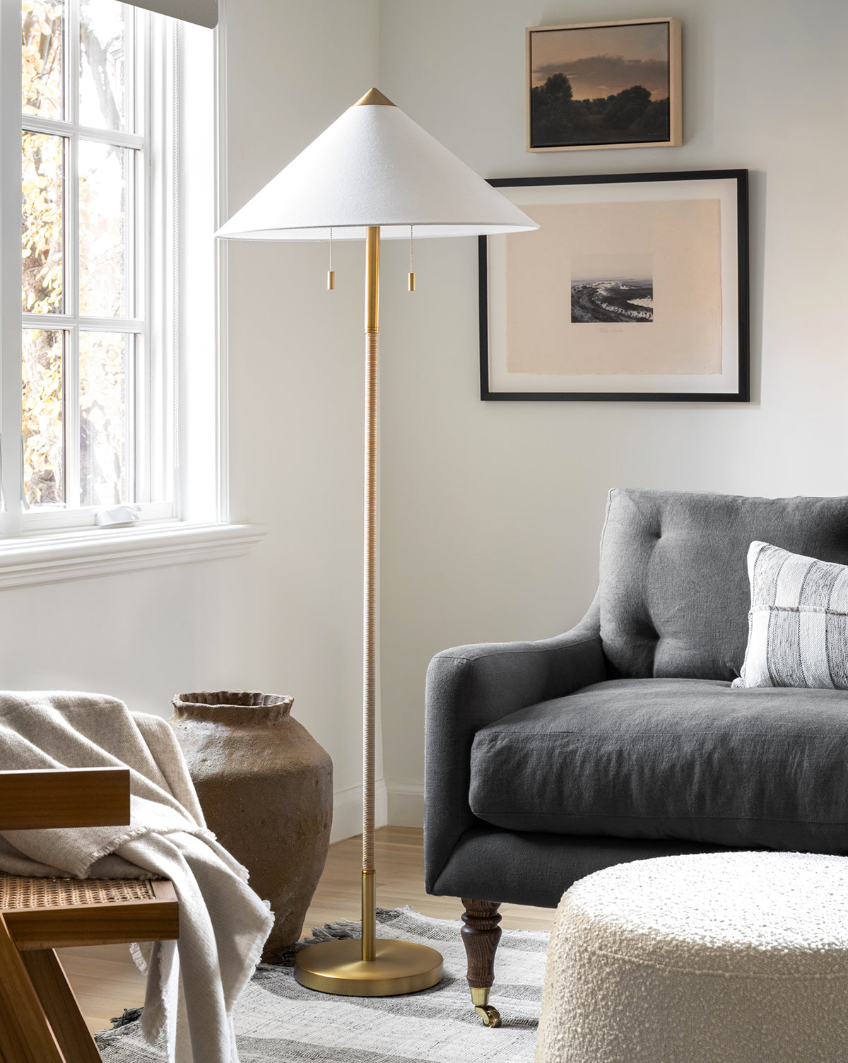 A modern living room features a gray sofa, floor lamp with white shade, framed The Grove by Bruce Brainard wall art, large window, textured vase, throw blanket, and round ottoman atop a light rug.