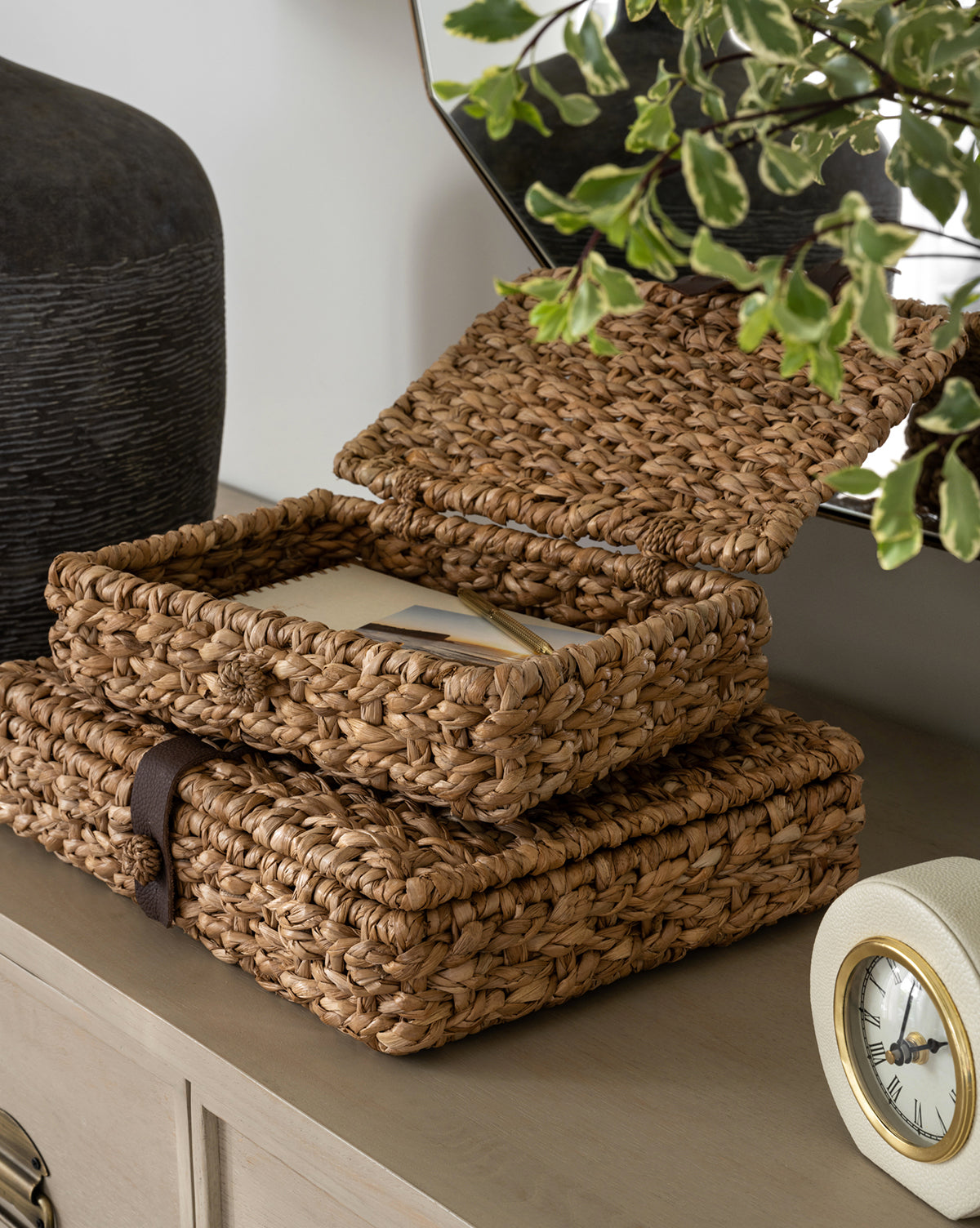 Two Corinne Woven Boxes are stacked on a wooden surface. The top woven box, lid open with envelopes inside, sits near a round clock, a leafy plant, and a decorative vase.