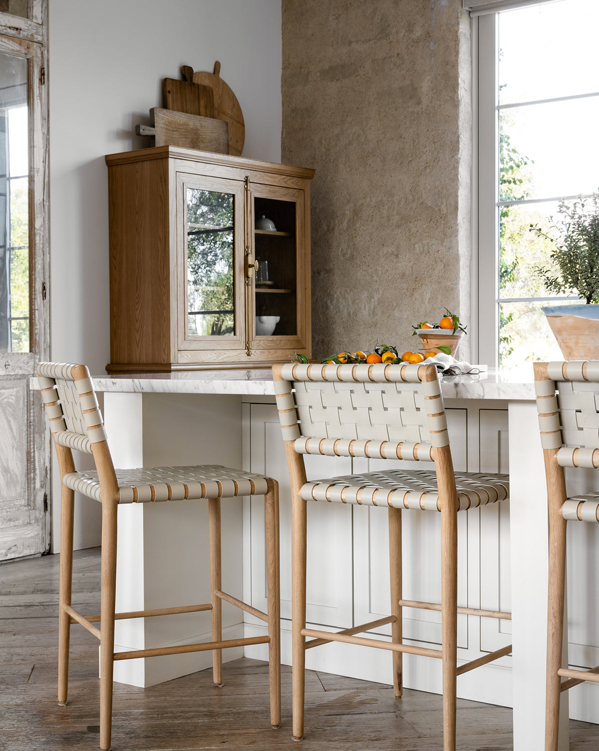 A bright, rustic kitchen features a marble island with three Greely Stools, a wooden cabinet, cutting boards, and a tray of oranges near a large window that fills the space with natural light by McGee & Co.