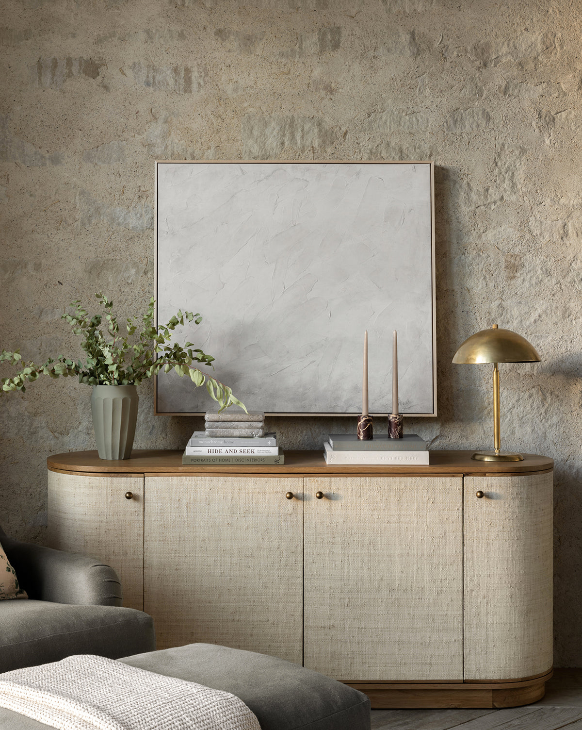 The Linu Woven Sideboard in neutral tones sits against a textured stone wall, adorned with a vase of greenery, stacked books, two candlesticks, and a brass lamp, by McGee & Co. Above it hangs a large square frame featuring abstract art.