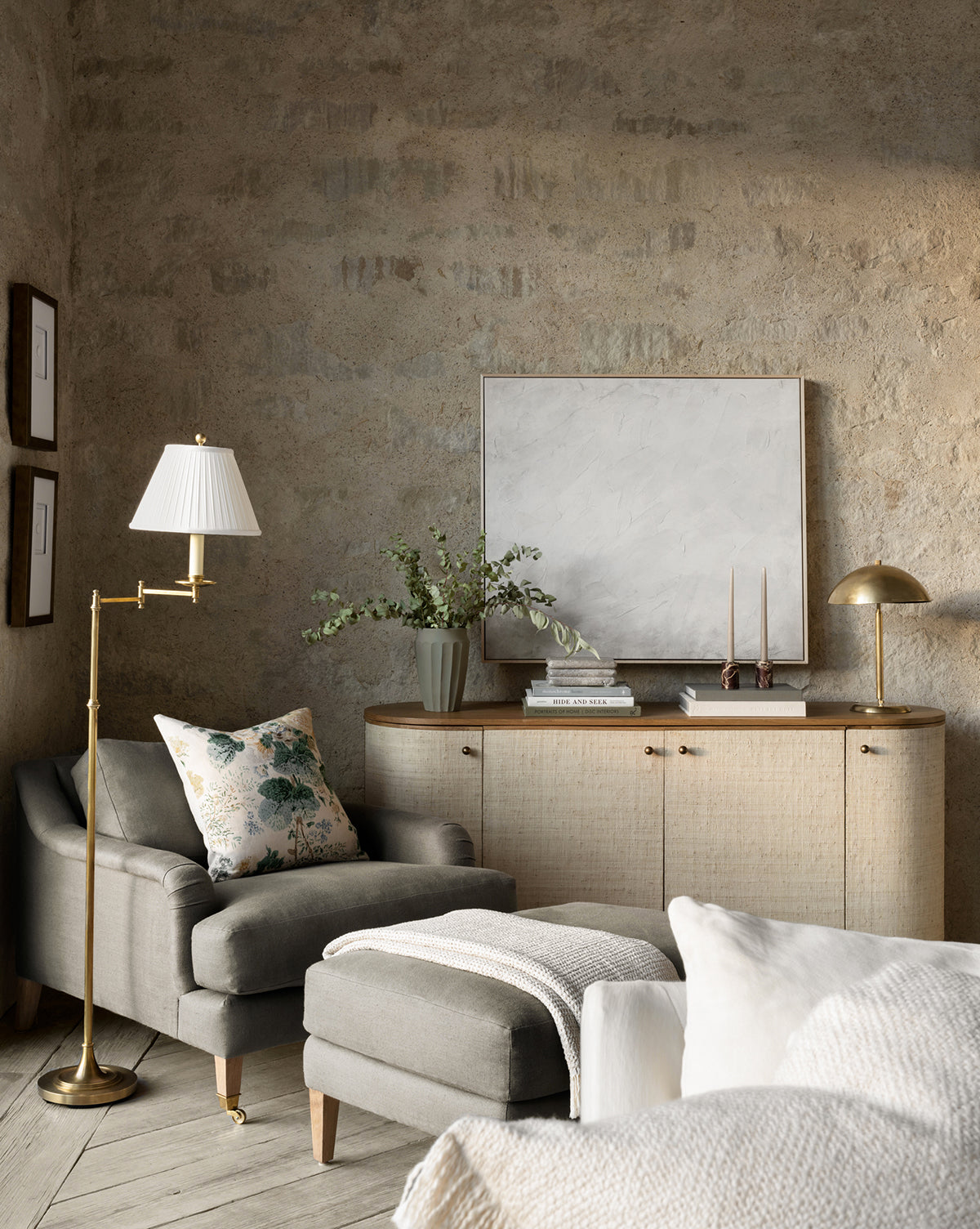 A cozy living room corner with a gray armchair, floral cushion, ottoman, cream console table, decor accents, framed art, textured stone wall in warm light, and the Dorchester Swing Arm Floor Lamp featuring a pleated silk shade, by McGee & Co.