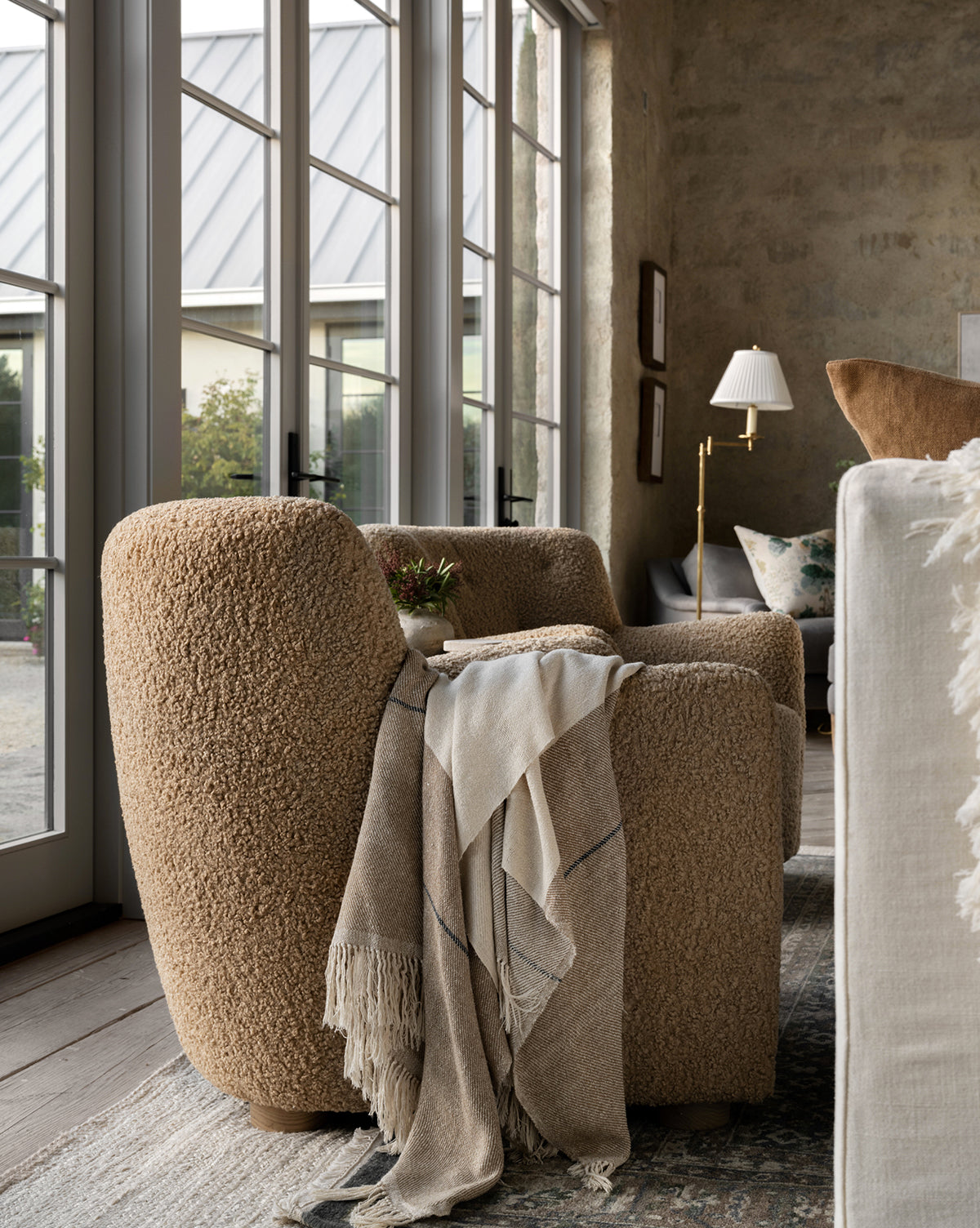 Two cozy, light brown Magda Lounge Chairs (Ready to Ship) face large glass doors. A fringed beige throw drapes over one chair, while a side table with a small plant sits between them in this sunlit, inviting living space by McGee & Co.
