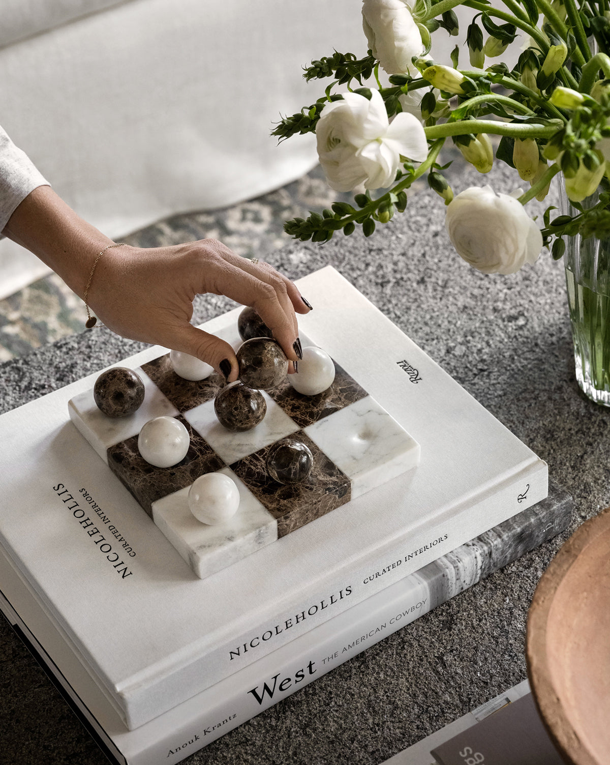 A hand moves a piece on the Marble Tic-Tac-Toe Board, set on stacked design books beside white flowers—a chic marble coffee table decor accent.