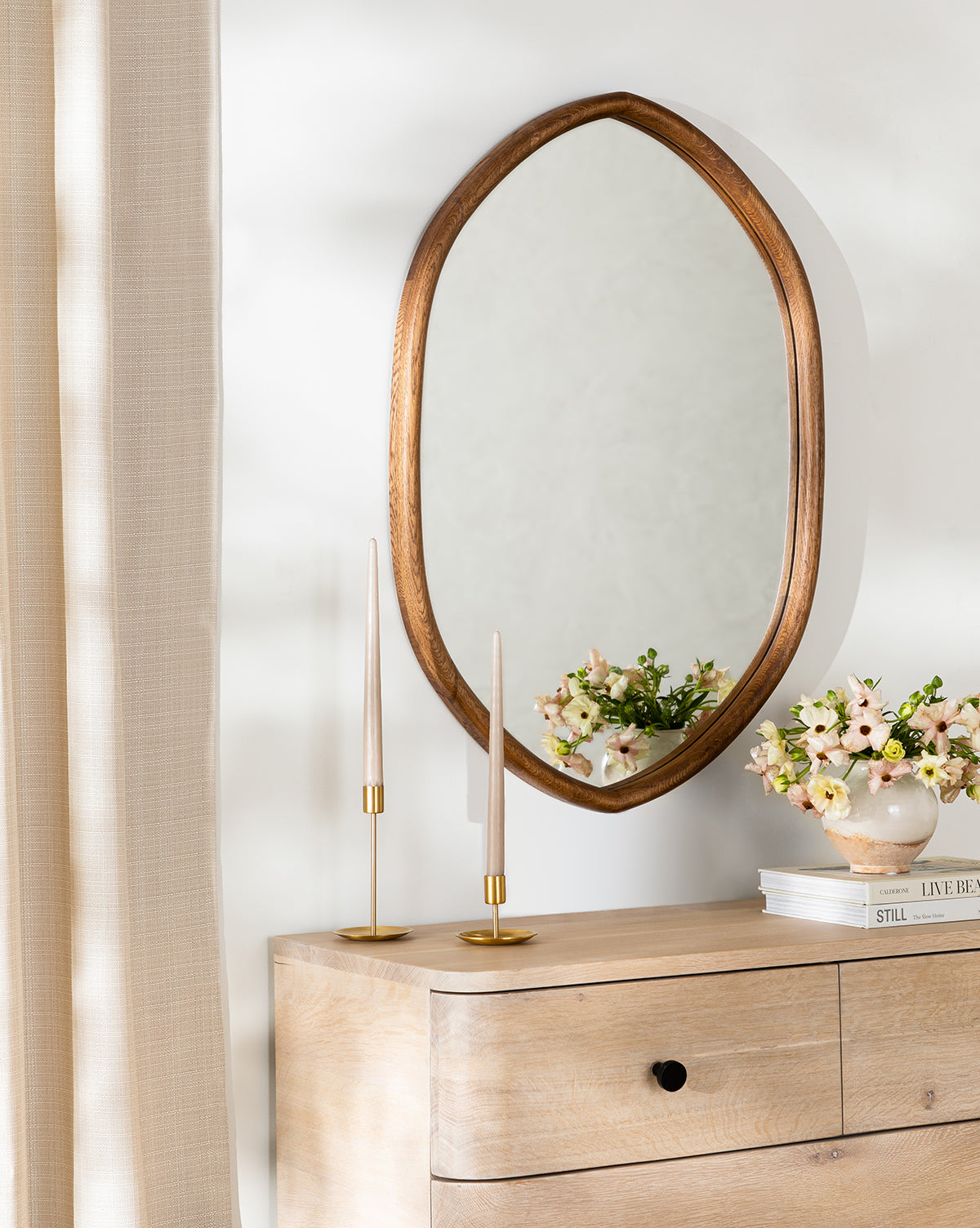 The Oslo Mirror with an oak frame hangs above a wooden dresser decorated with gold candlesticks, a pink flower vase, and a bowl, beside beige curtains and against a white wall, by McGee & Co.