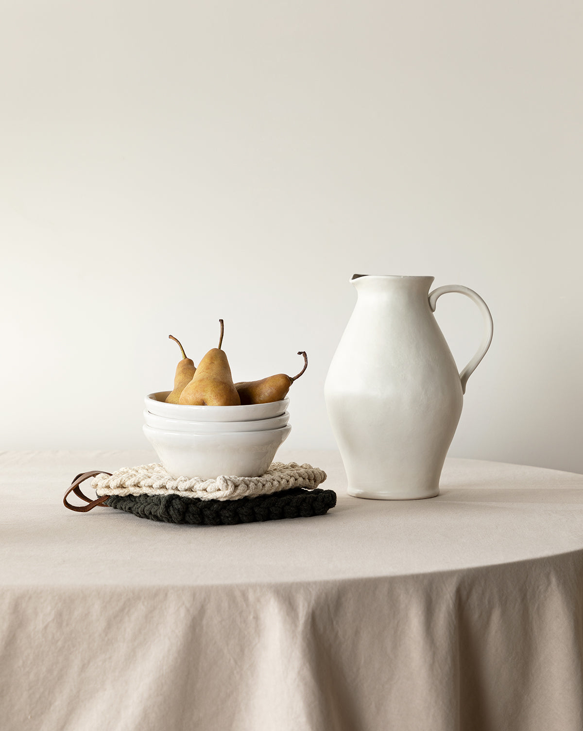 A still life shows the White Ceramic Pitcher with a matte finish beside a white bowl of pears on a beige tablecloth, with knitted trivets under the bowl against a plain light backdrop, by McGee & Co.
