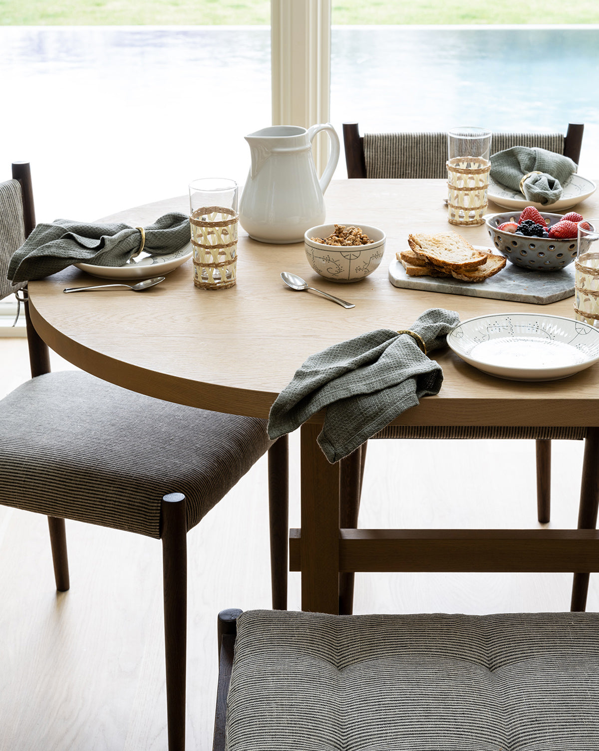 The Giselle Oval Dining Table is set for breakfast with plates, bowls of cereal, a bowl of berries, toast on a plate, a white pitcher, glasses of water, gray napkins, and cushioned chairs arranged around it by McGee & Co.
