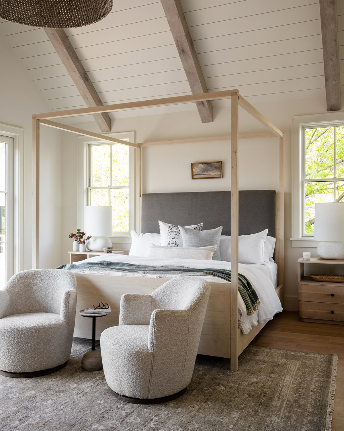 A modern bedroom features a wooden canopy bed with a gray upholstered headboard, white bedding, and large windows. Two Gulliver Swivel Chairs in boucle fabric and a small round side table complete the inviting space.