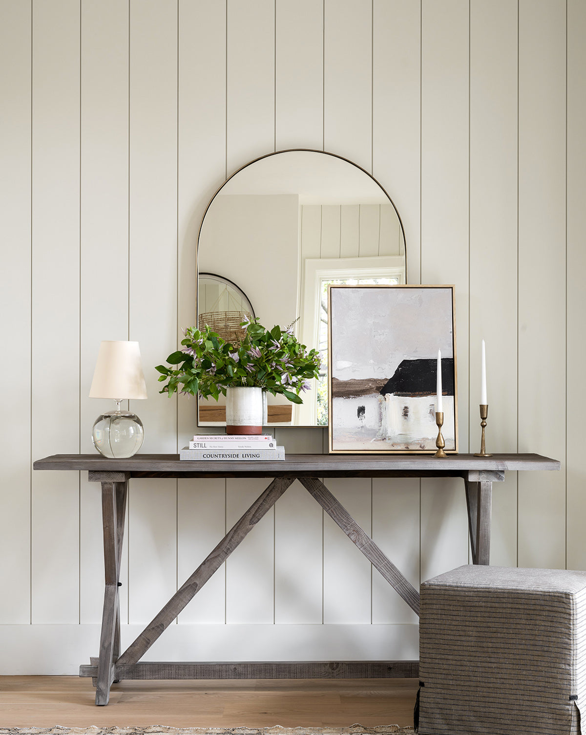 A wooden console table with décor sits against a white paneled wall, the Wylie Arched Mirror with shatterproof glass hanging above and a gray ottoman nearby, by McGee & Co.