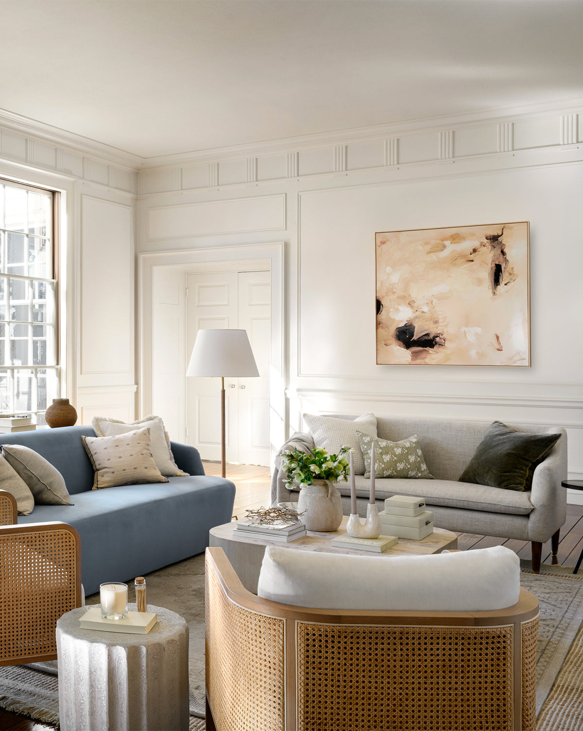 Bright, elegant living room with a blue sofa, gray loveseat, rattan chairs, a round side table, and the Warm Abstract wall art. Large window fills space with light; decor includes neutral pillows, plants, and candles.