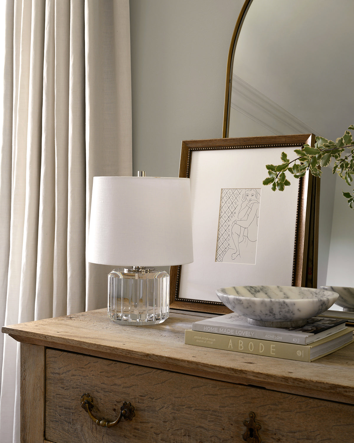 A wooden dresser holds a crystal table lamp with a white shade, the Simple Sketched Woman artwork in a traditional frame, a marble bowl, two books, and a leafy branch. Light curtains and a curved mirror are in the background.