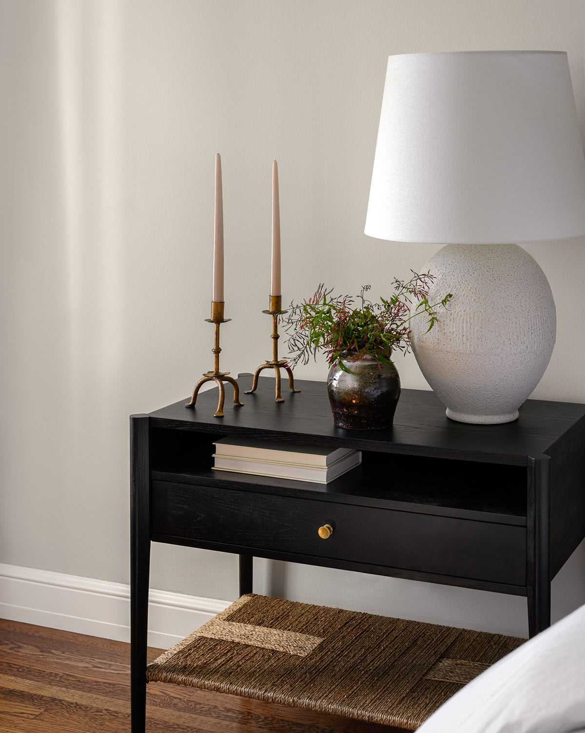 The Dana Nightstand in a black finish features a drawer and displays a white lamp, two brass candlesticks with tapered candles, a small vase with greenery, and a closed book, by McGee & Co. A woven seagrass shelf below adds texture to the light wall backdrop.