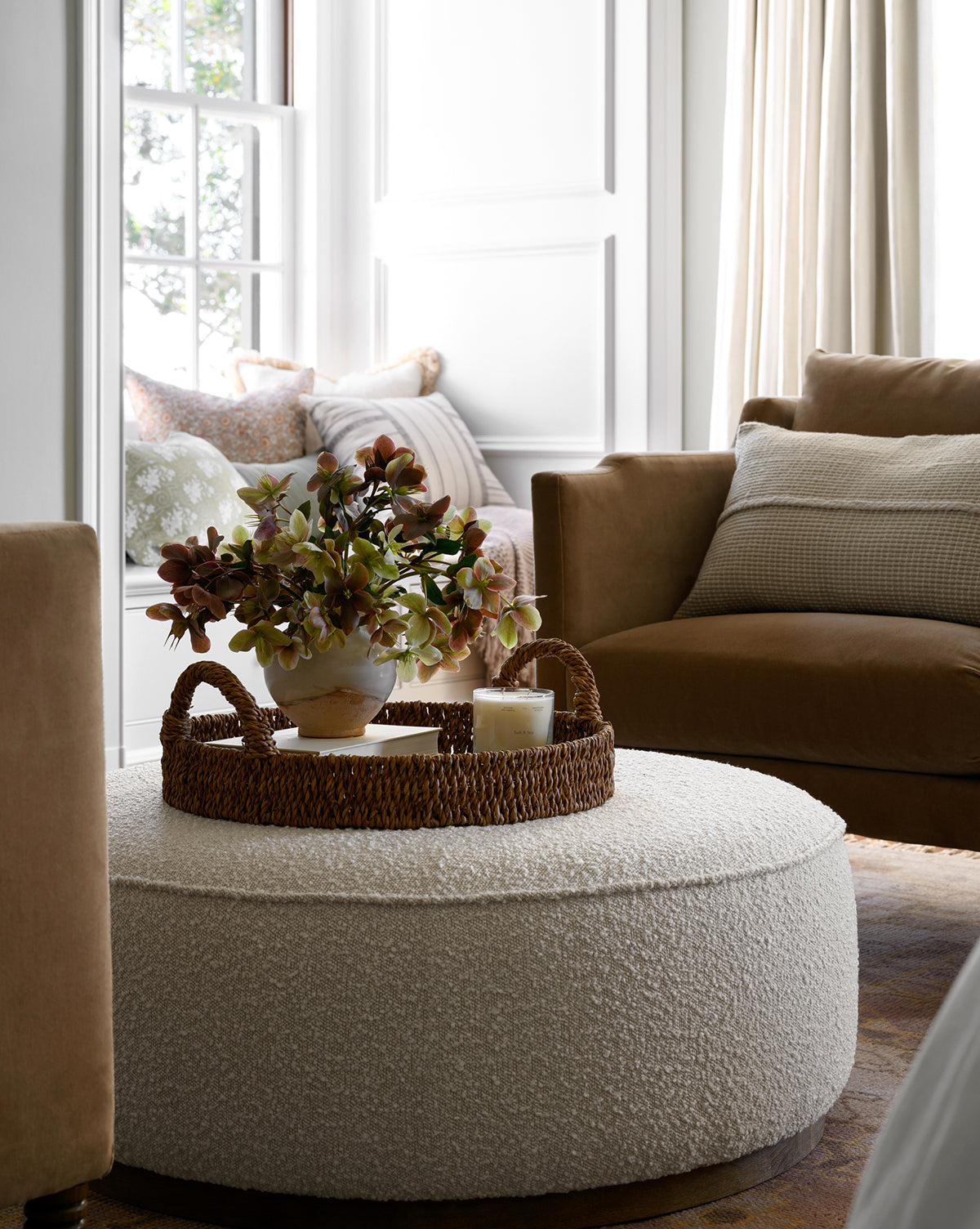 A cozy living room showcases the Dawson Ottoman with a fabric cushion and wood base topped by a woven tray, floral arrangement, and candle, set beside a brown sofa with soft pillows near a sunlit window, by McGee & Co.