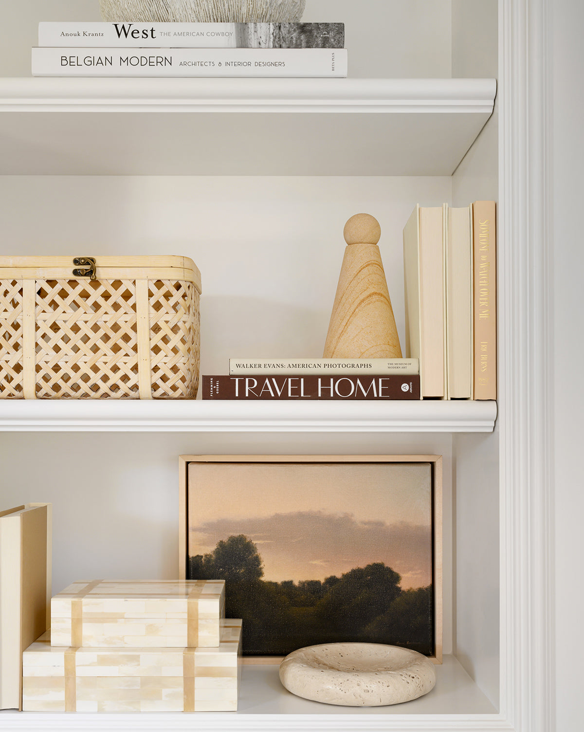 A neatly styled white shelf displays neutral books, a wicker storage basket, a wooden figure, stacked decorative boxes including the Paige Bone Box, a round stone accent, and a framed landscape painting below.