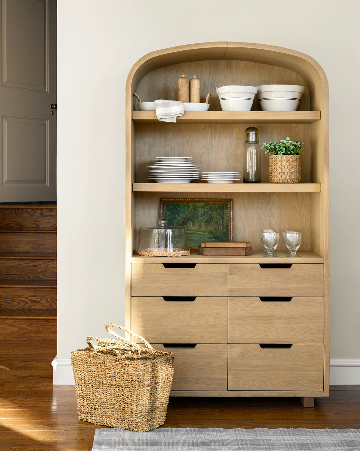 A Burnham Cabinet in light oak with six drawers and two open shelves displays white dishes, glassware, a framed painting, and decor. A woven basket is placed on the floor next to it, set against wood floors and neutral walls, by McGee & Co.